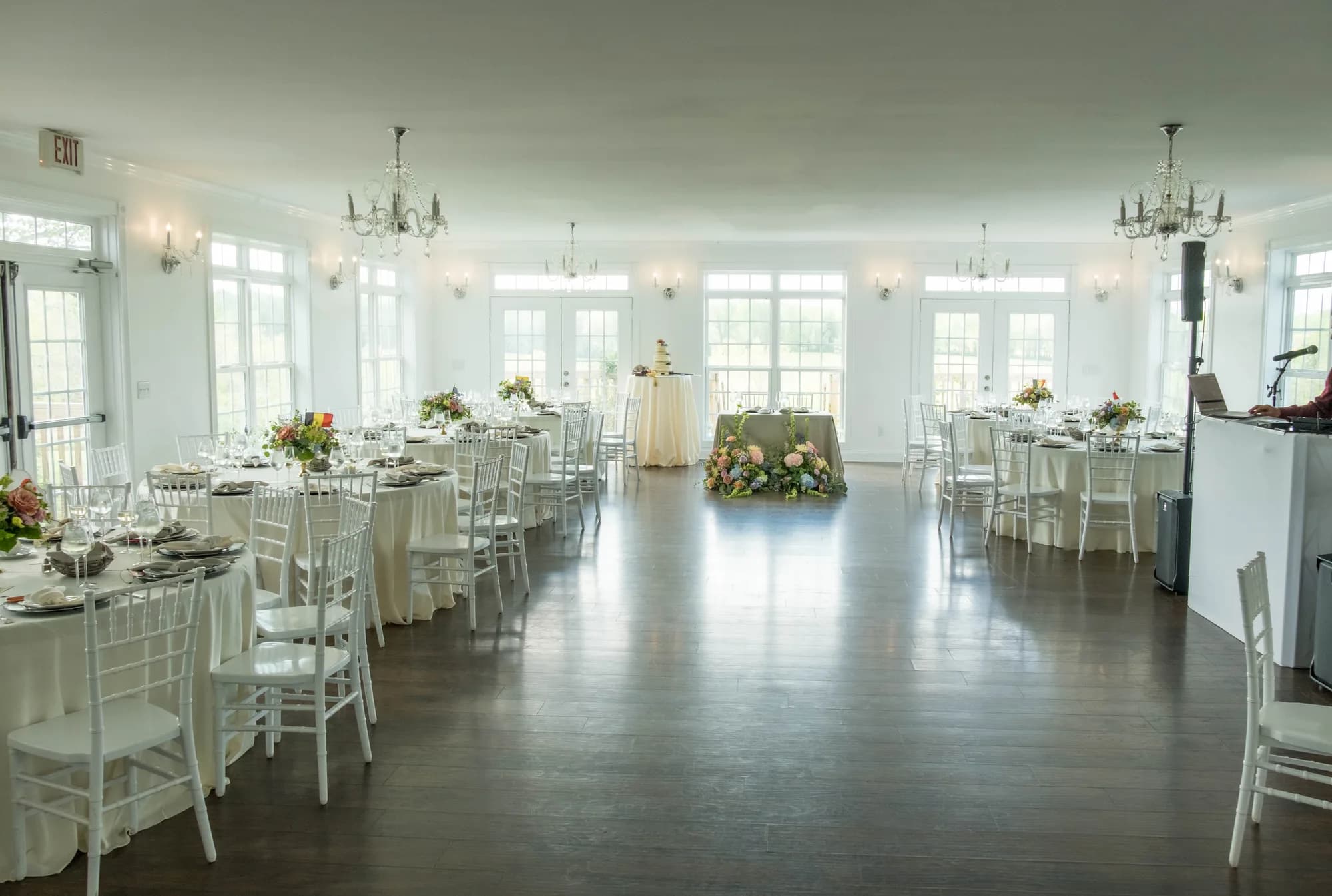 Sunlit Rixey Manor ballroom set for a wedding reception with white chiavari chairs, floral centerpieces, and a cake table