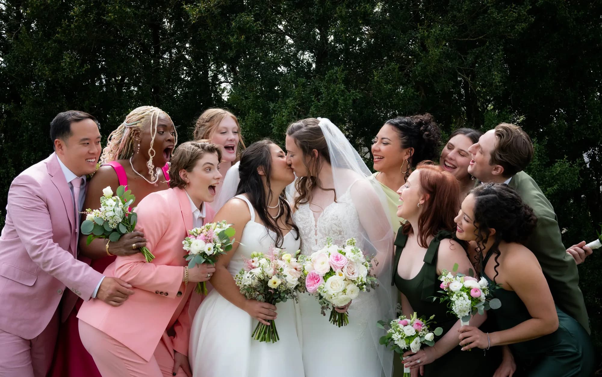 Two brides share a kiss surrounded by a joyful wedding party erupting in laughter and cheers outdoors