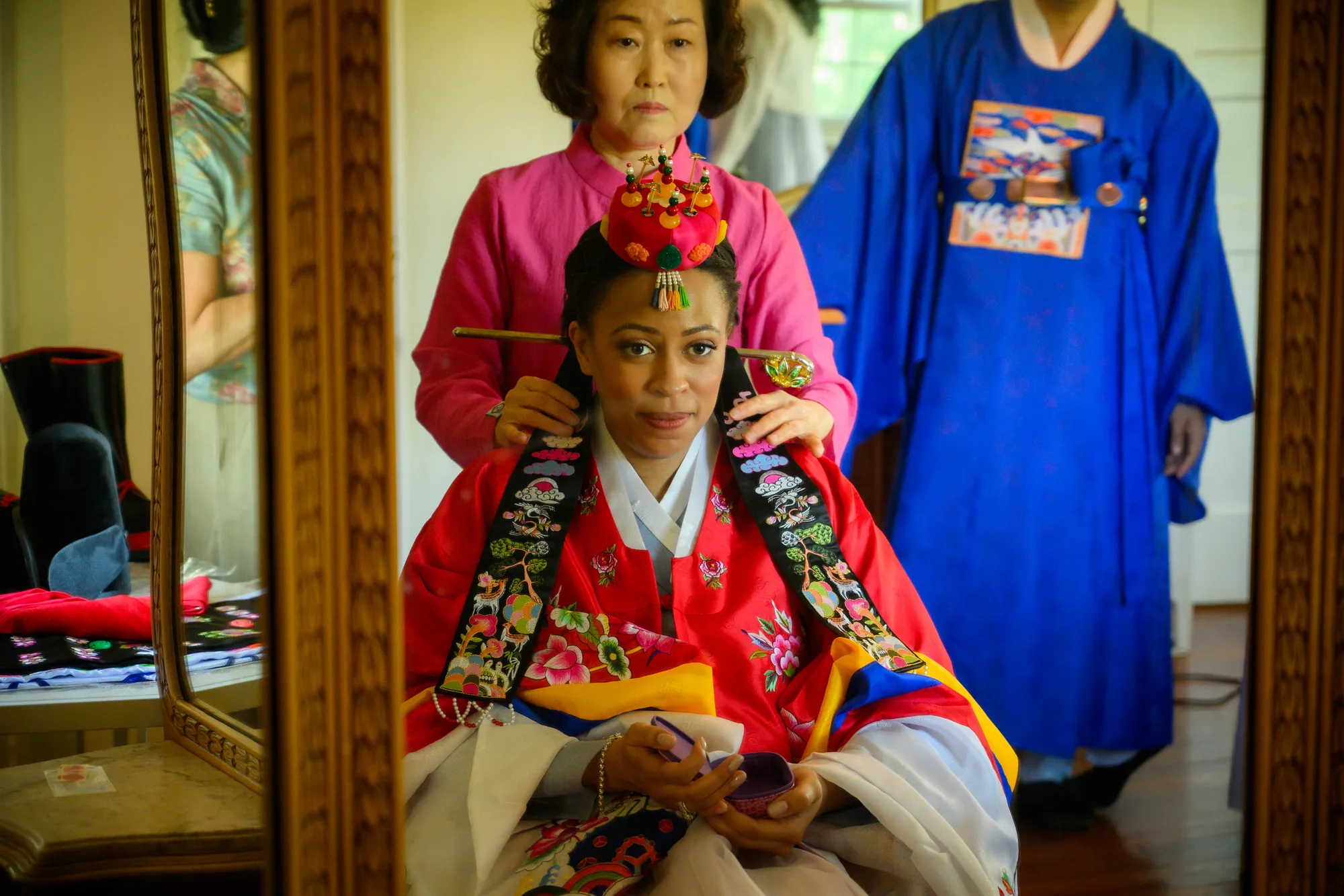 Bride in red Korean hanbok has traditional hair ornament placed by attendant before wedding ceremony