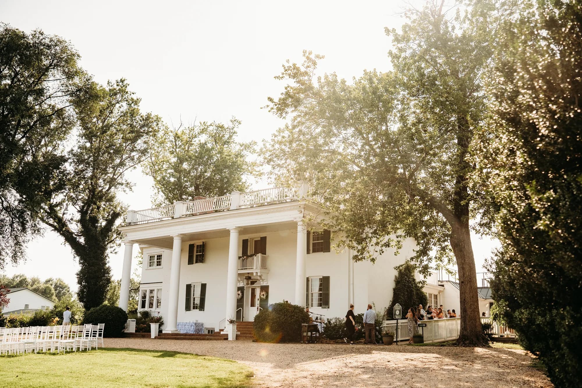 Sunlit exterior of Rixey Manor estate with white columns, mature trees, and guests gathering on the grounds