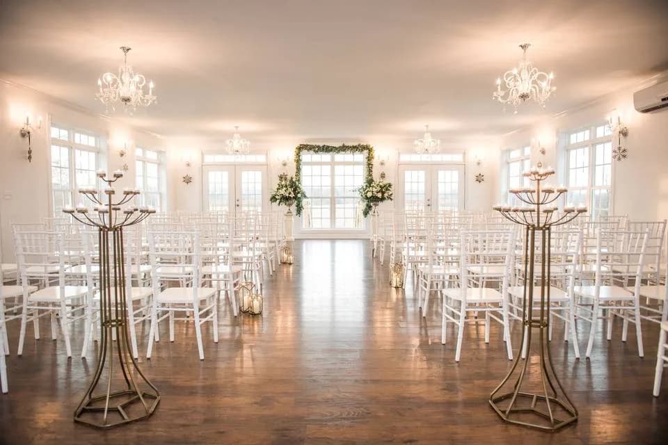 The Rixey Manor ballroom set for a ceremony with white chiavari chairs, gold candelabras, and a greenery arch in front of the French doors.