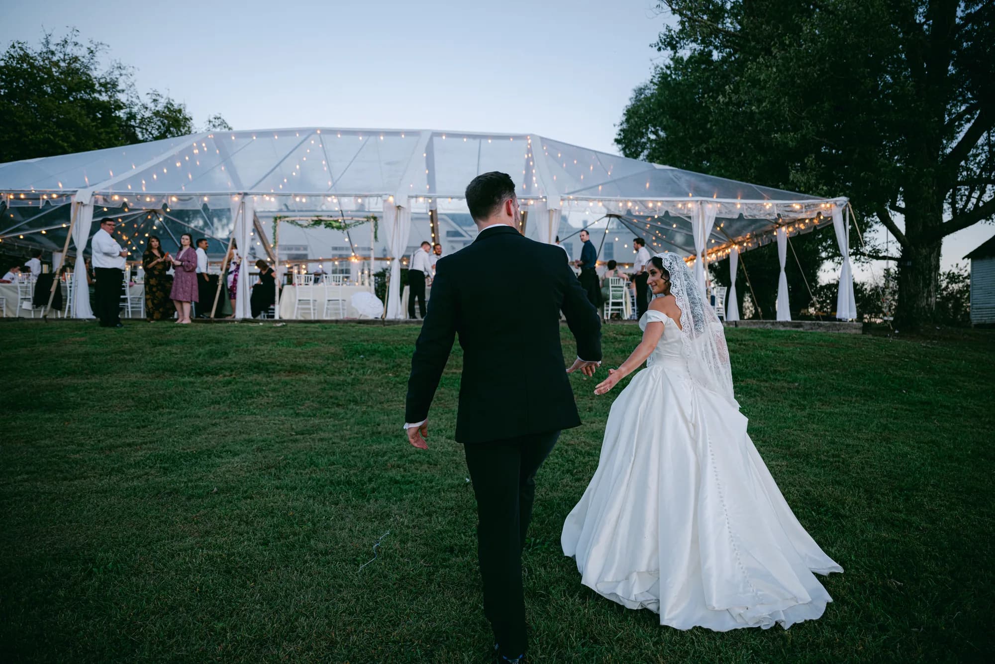Bride and groom walk hand-in-hand toward a glowing tent reception on lush Rixey Manor grounds at dusk
