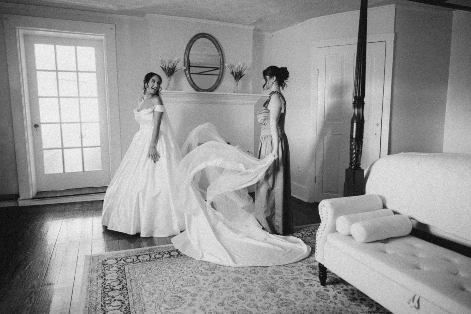 Bride's veil billows dramatically as attendant assists in a sunlit historic room at Rixey Manor