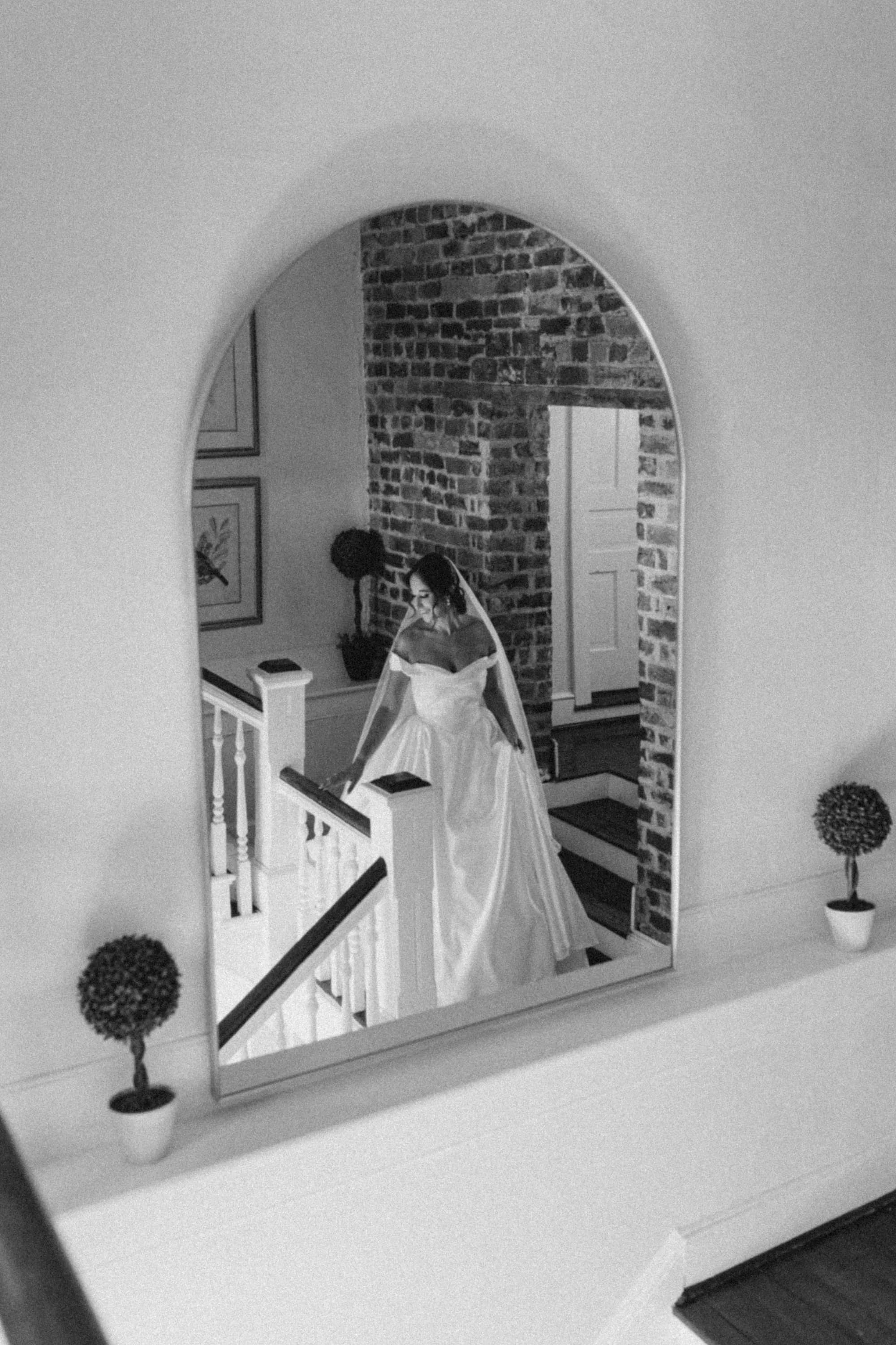 Bride in wedding gown reflected in arched mirror on staircase landing with exposed brick wall at Rixey Manor