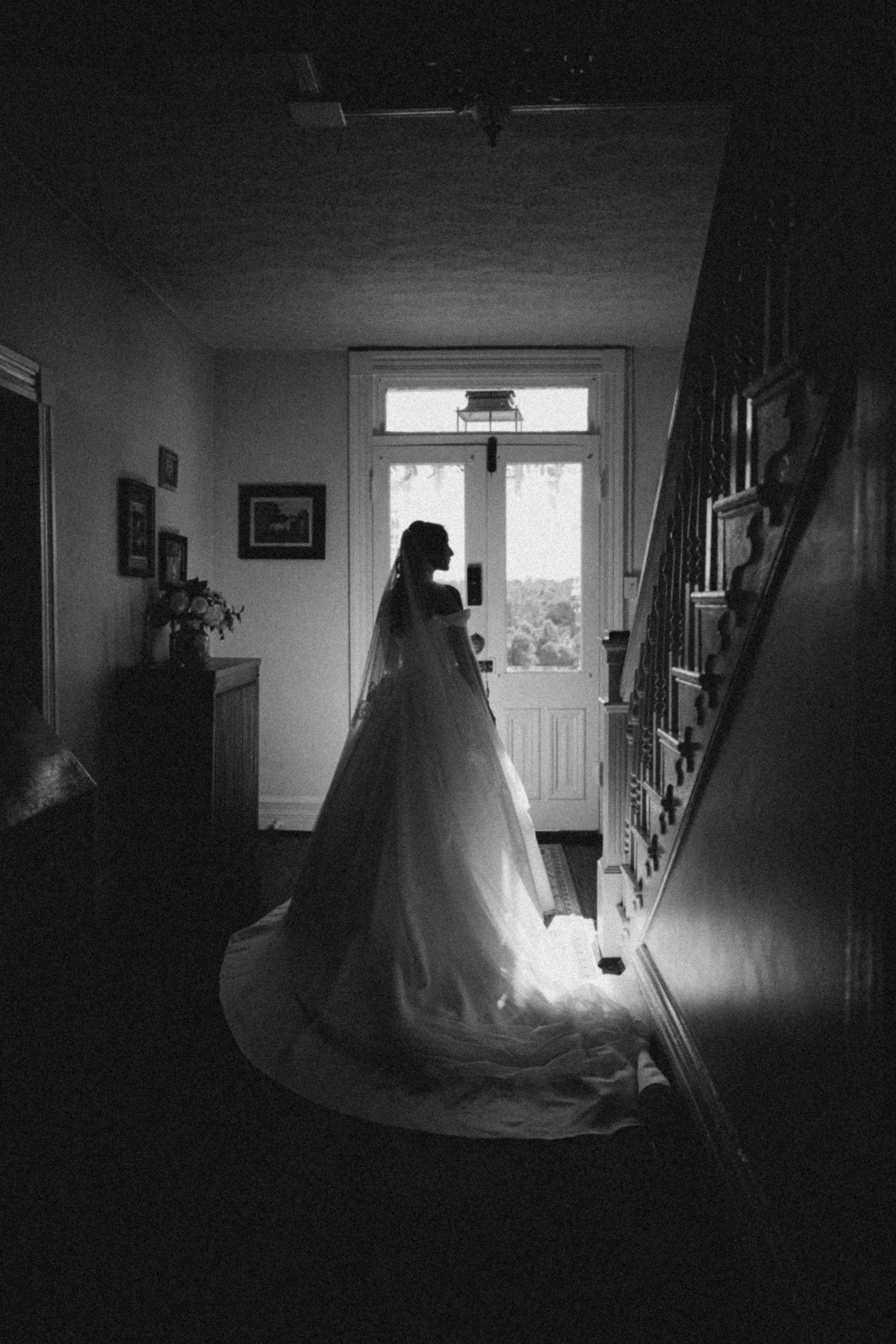 Bride in full gown and veil silhouetted against a sunlit front door inside a historic manor foyer