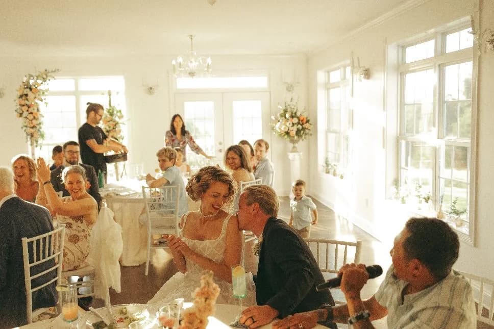 Bride laughing with guests at a sunlit reception inside an elegant white manor dining room