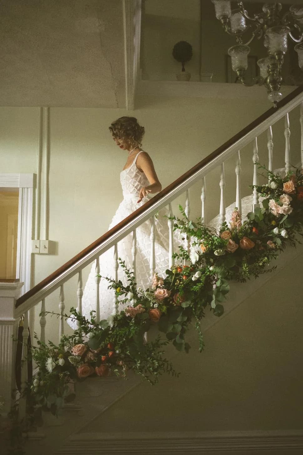 Bride descending flower-draped staircase at Rixey Manor in a lace gown, chandelier above