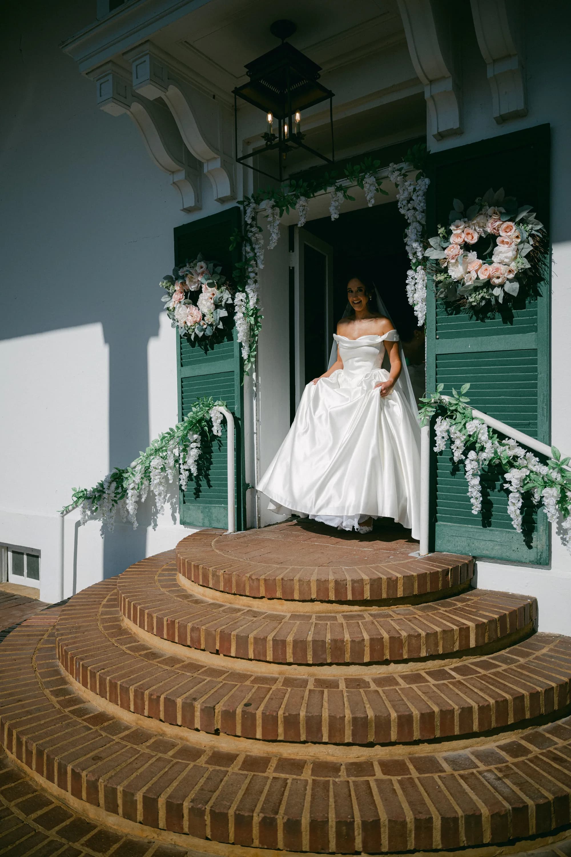 Bride in off-shoulder gown standing in flower-adorned doorway atop curved brick steps at Rixey Manor