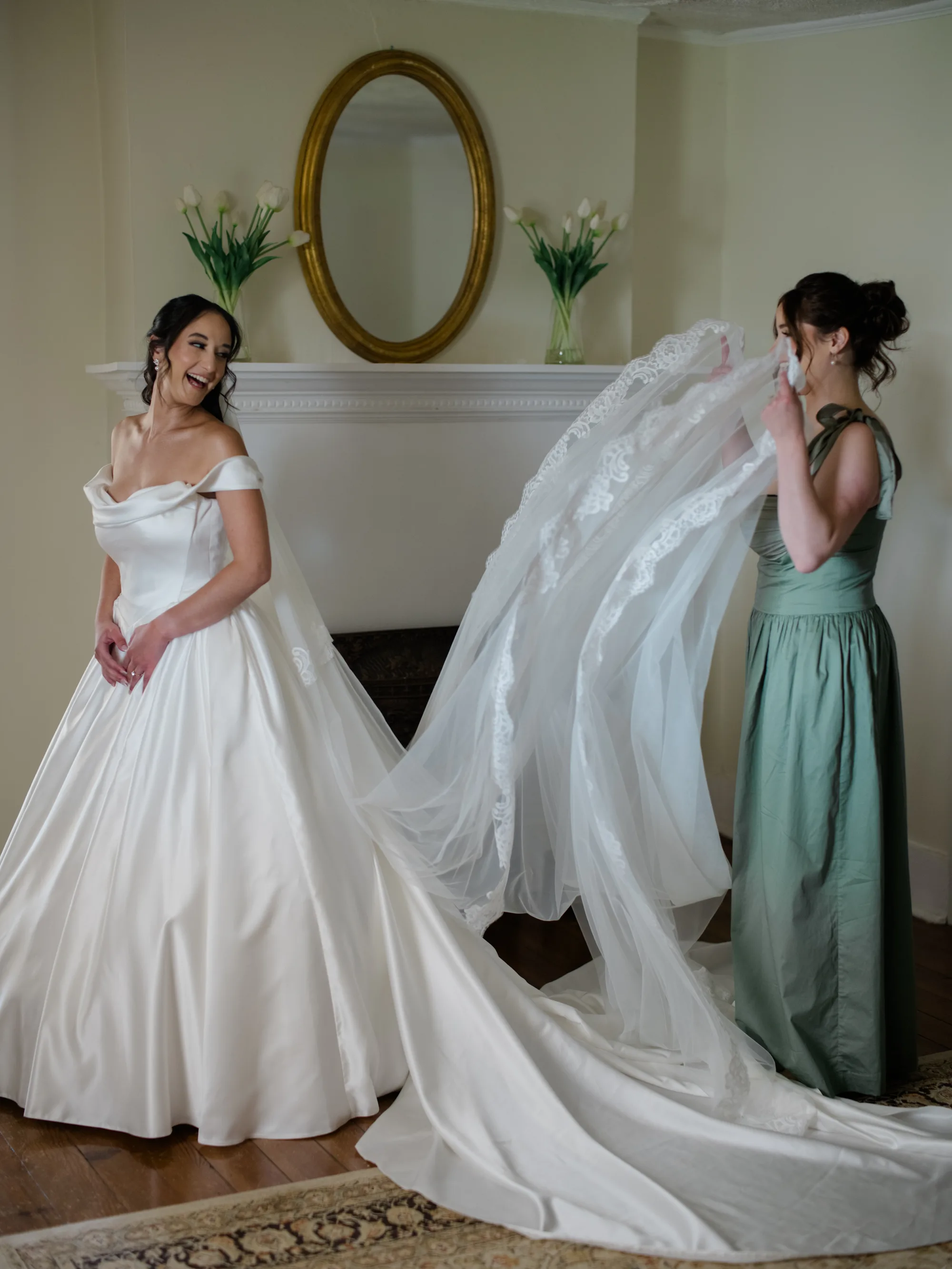 Laughing bride in off-shoulder gown as bridesmaid arranges lace veil near a fireplace