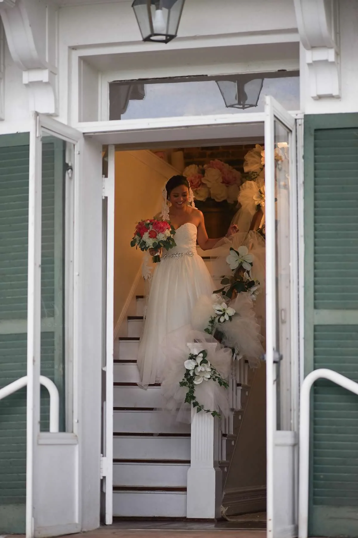 Smiling bride in white gown descends flower-adorned staircase through grand double doors at Rixey Manor