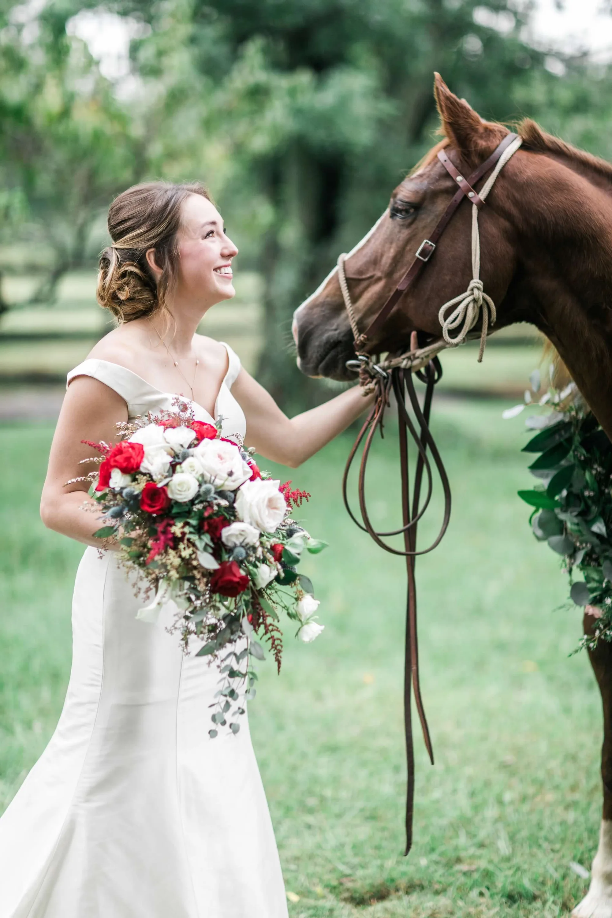 Smiling bride in white gown holds red and white bouquet while petting a chestnut horse on lush green grounds