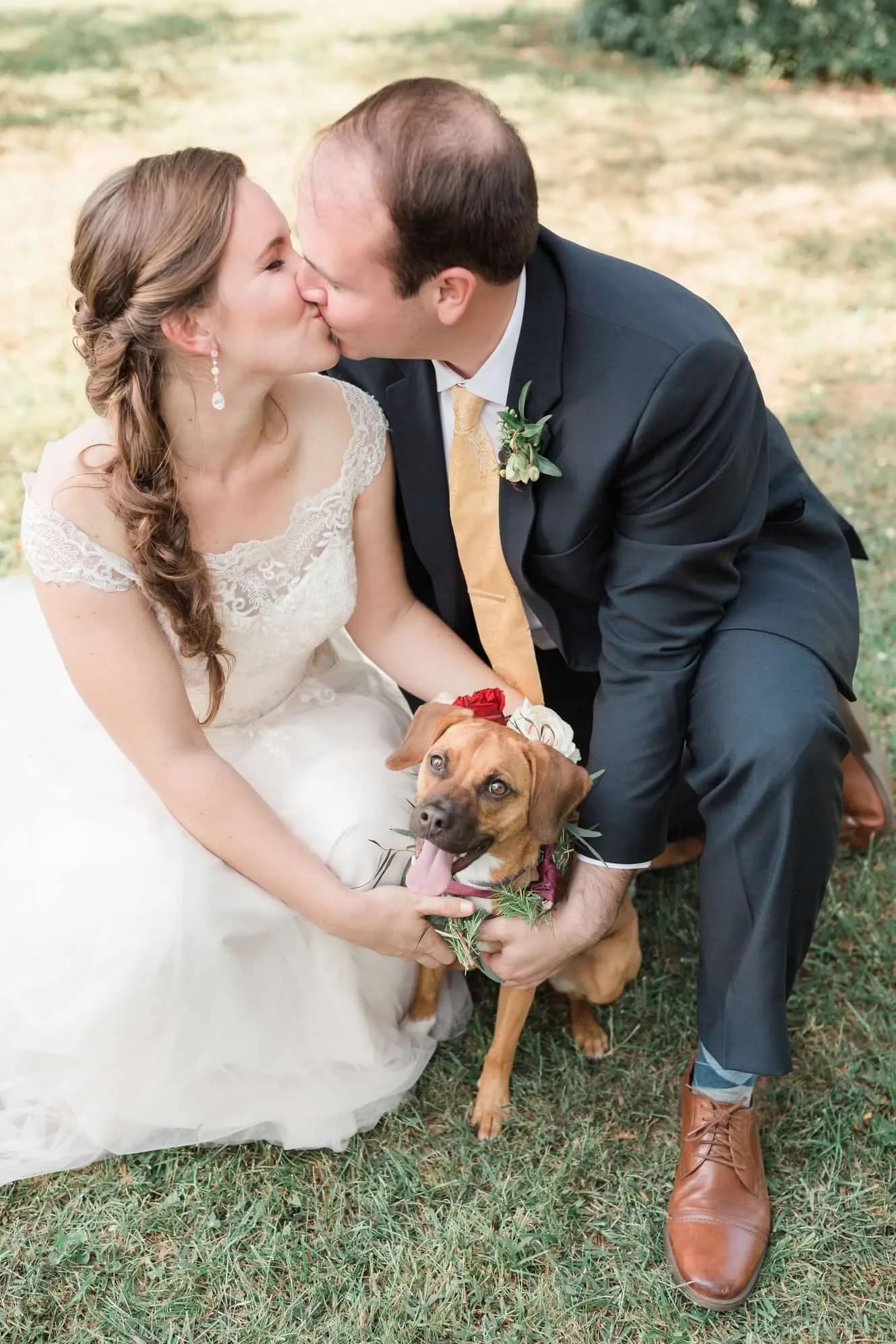 Bride and groom kiss outdoors while holding their flower-crowned dog on their wedding day
