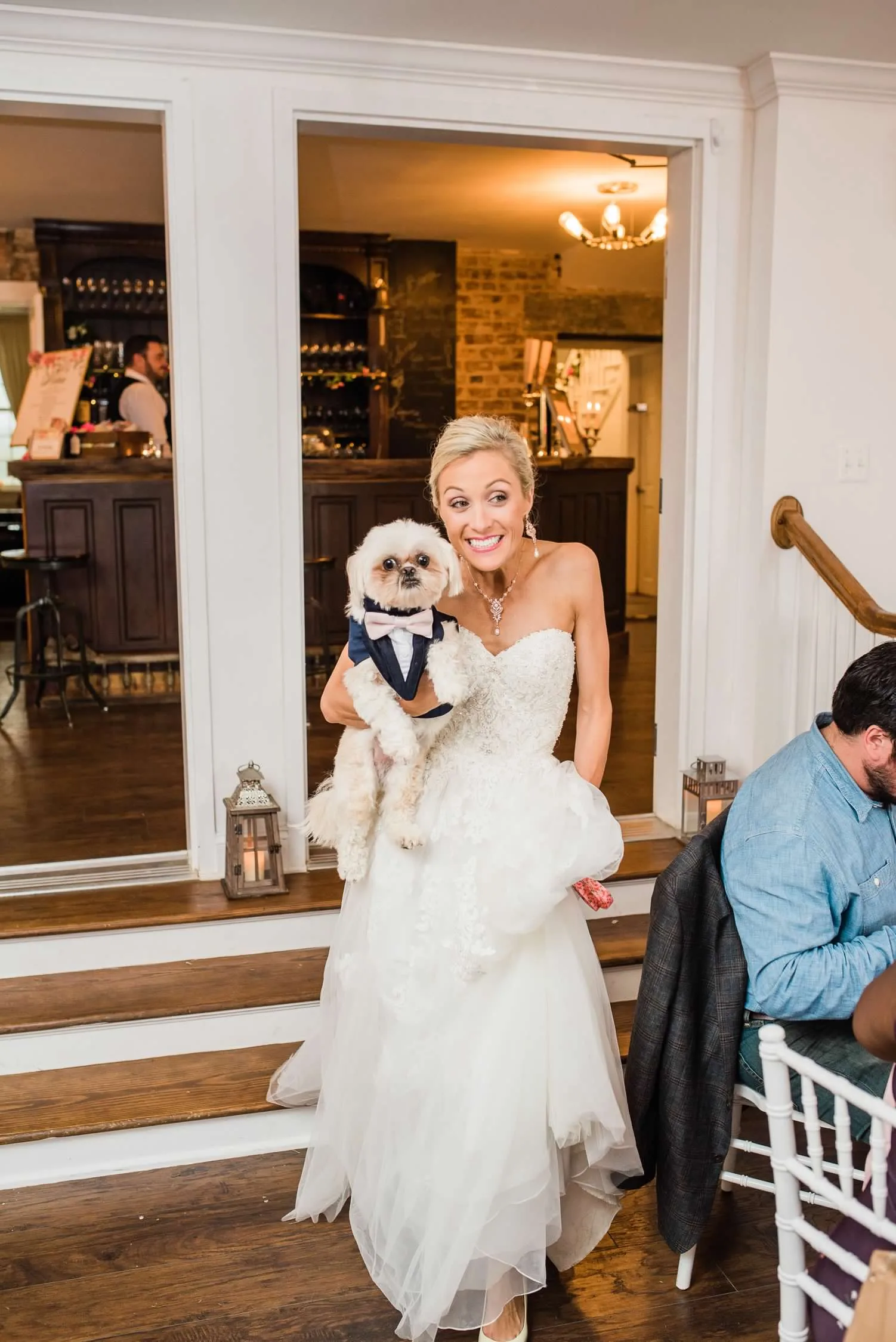 Smiling bride in strapless gown holds small white dog dressed in navy tuxedo at indoor wedding reception