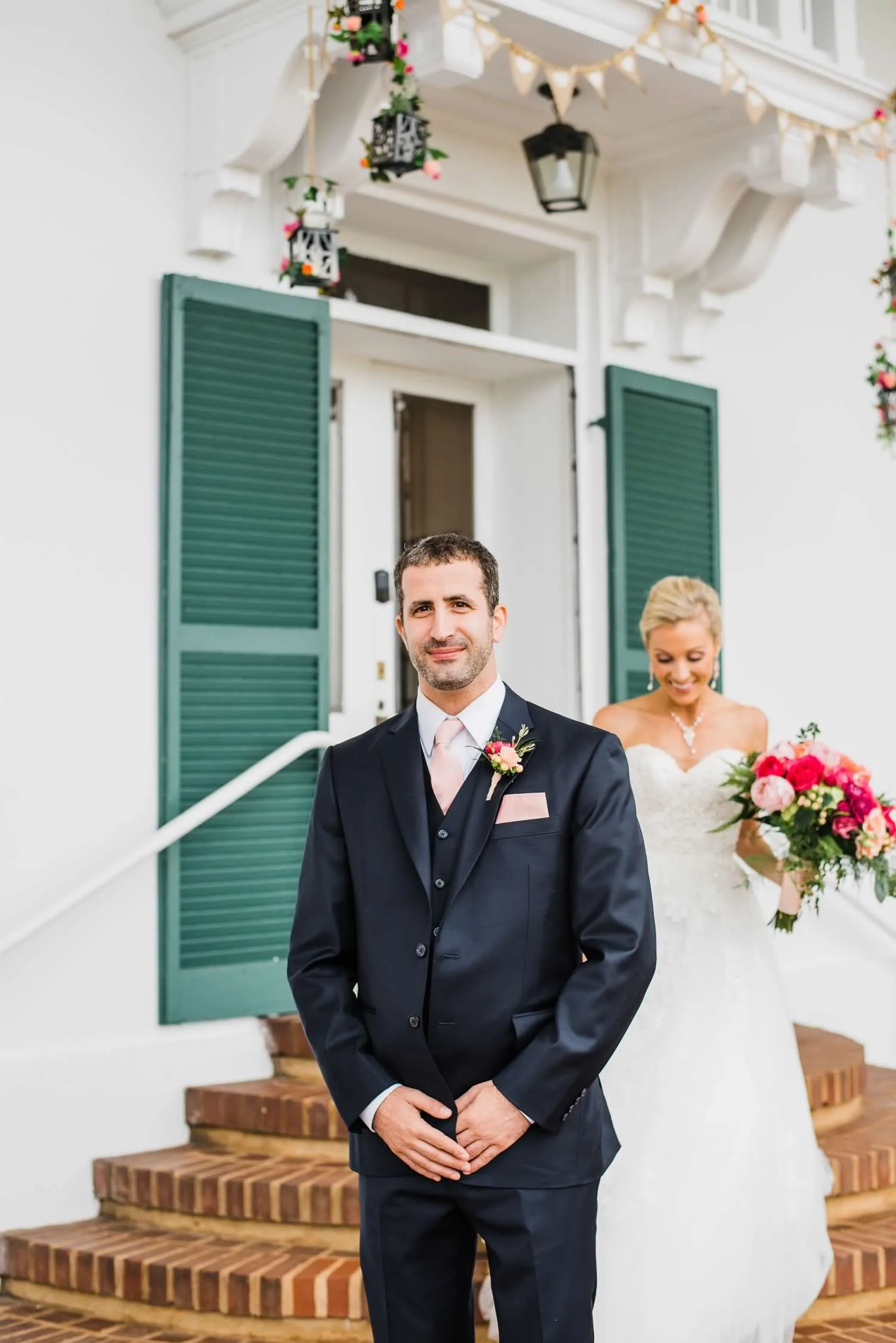 Groom in navy suit and bride in white dress pose for portraits outside white manor with green shutters.