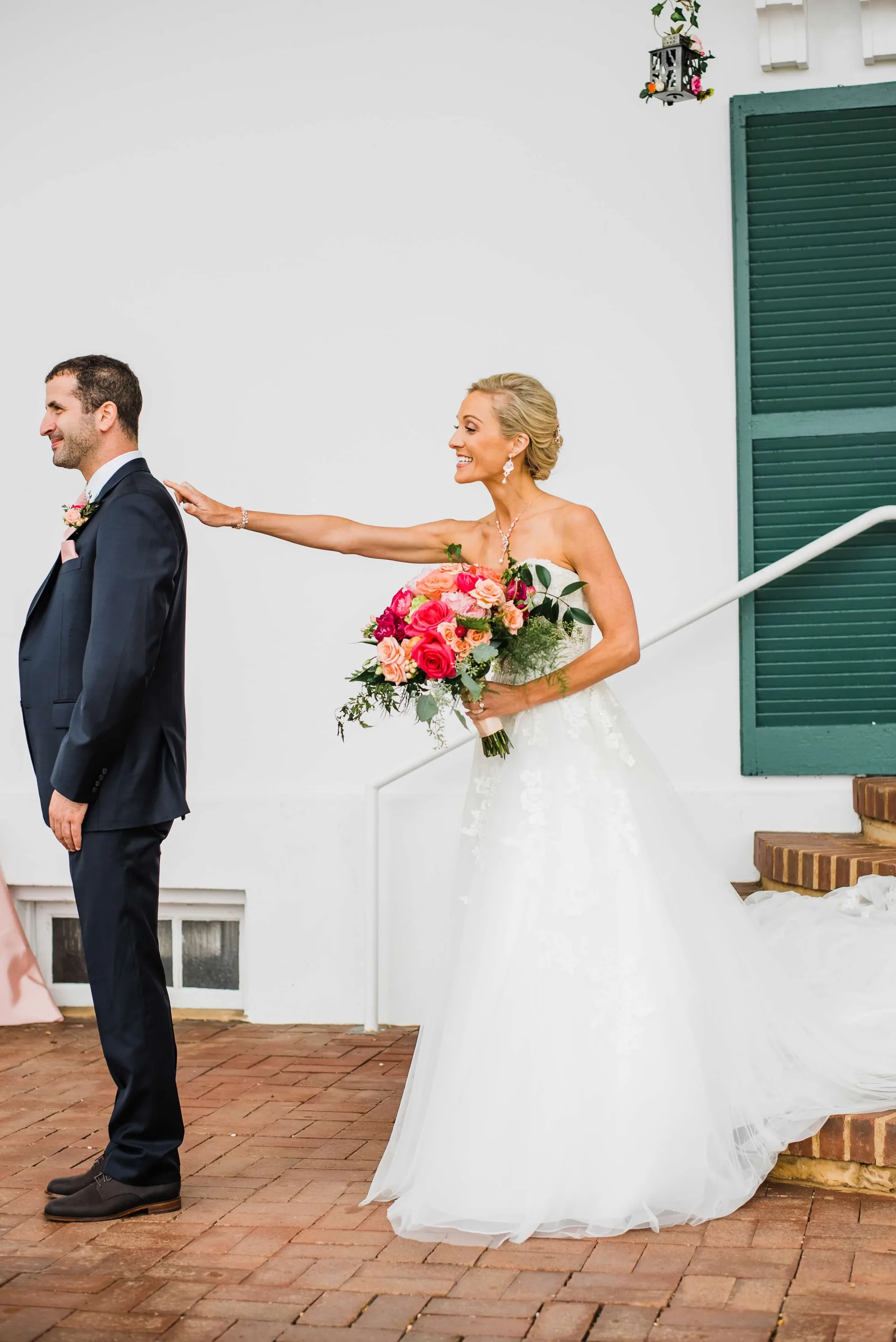 Bride taps groom's shoulder during first look on brick terrace, holding vibrant pink bouquet
