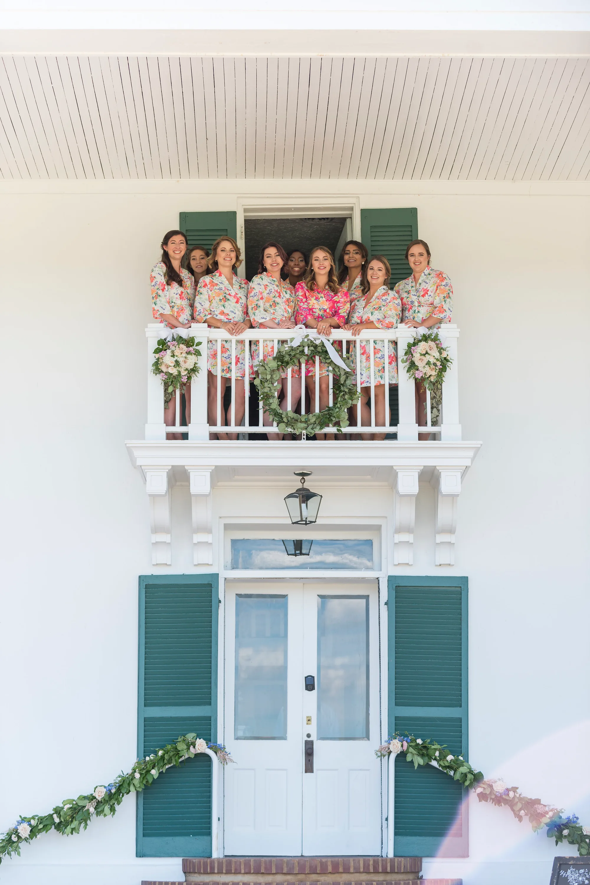 Bridesmaids in floral robes smile from a white balcony at Rixey Manor, adorned with a greenery wreath