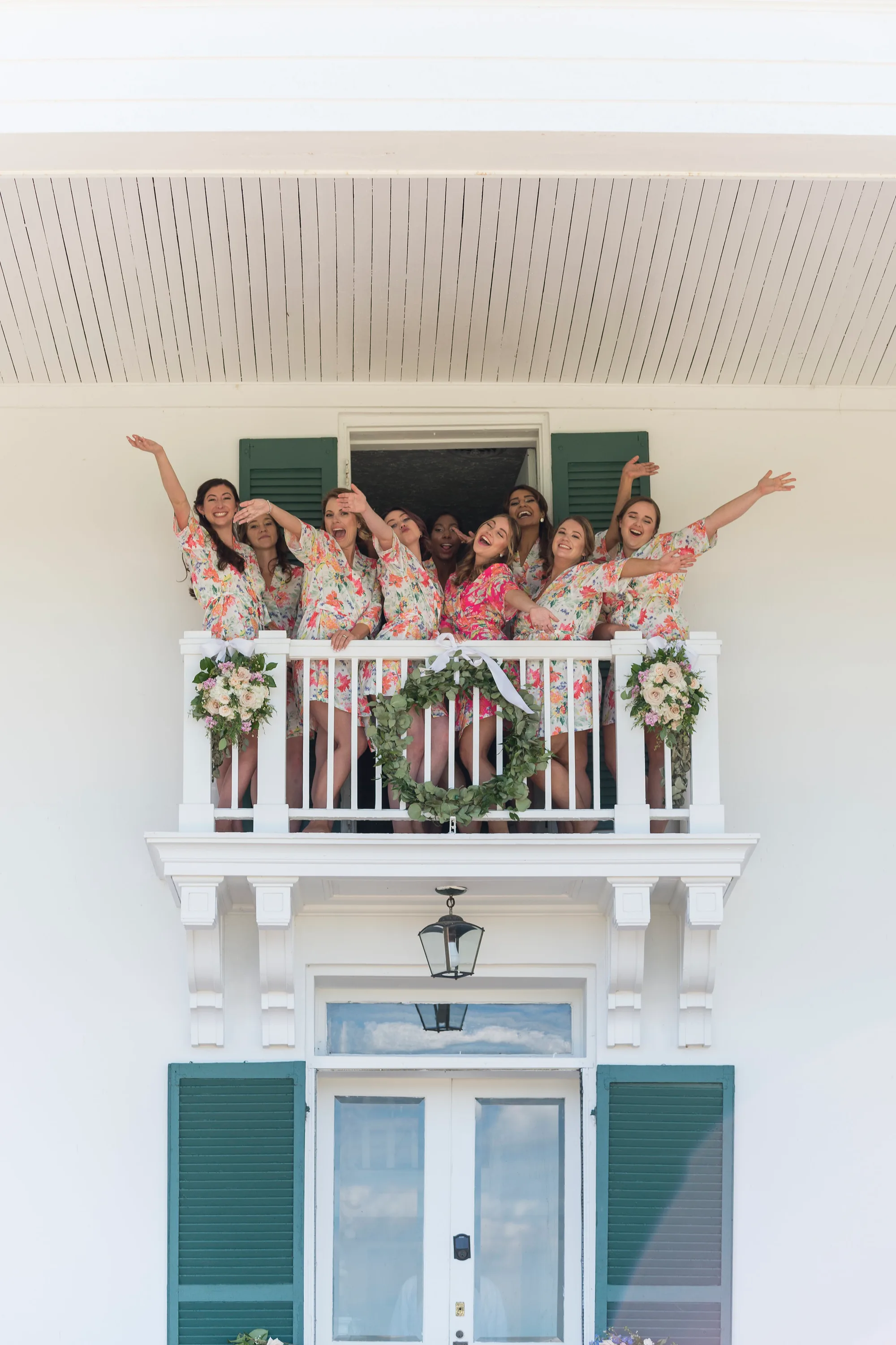 Bridesmaids in floral robes cheering from a white balcony adorned with greenery at Rixey Manor