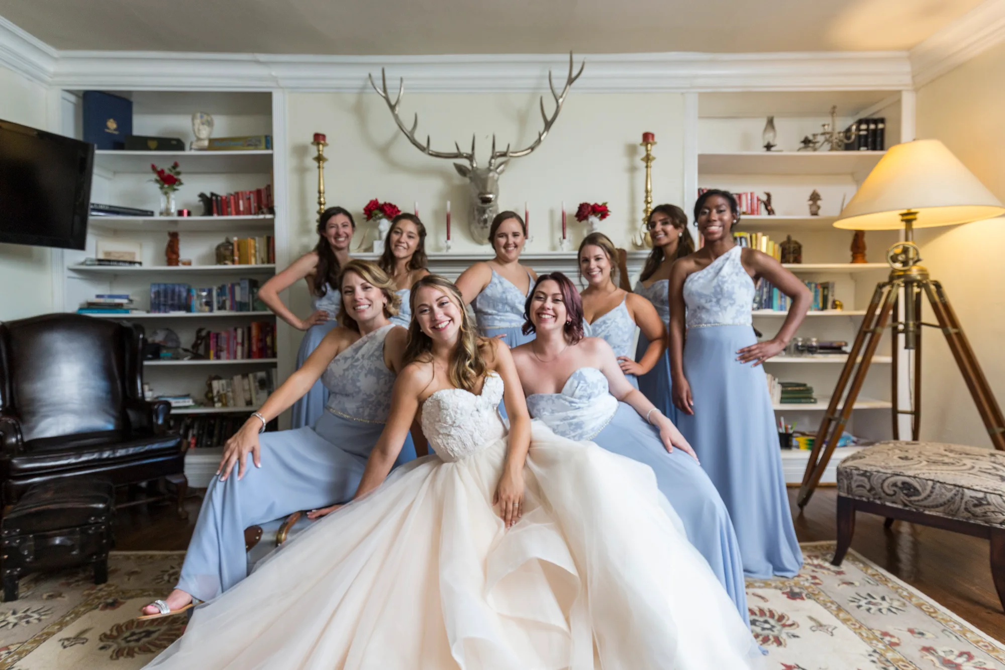 Bride surrounded by eight bridesmaids in blue gowns laughing together inside a Rixey Manor sitting room