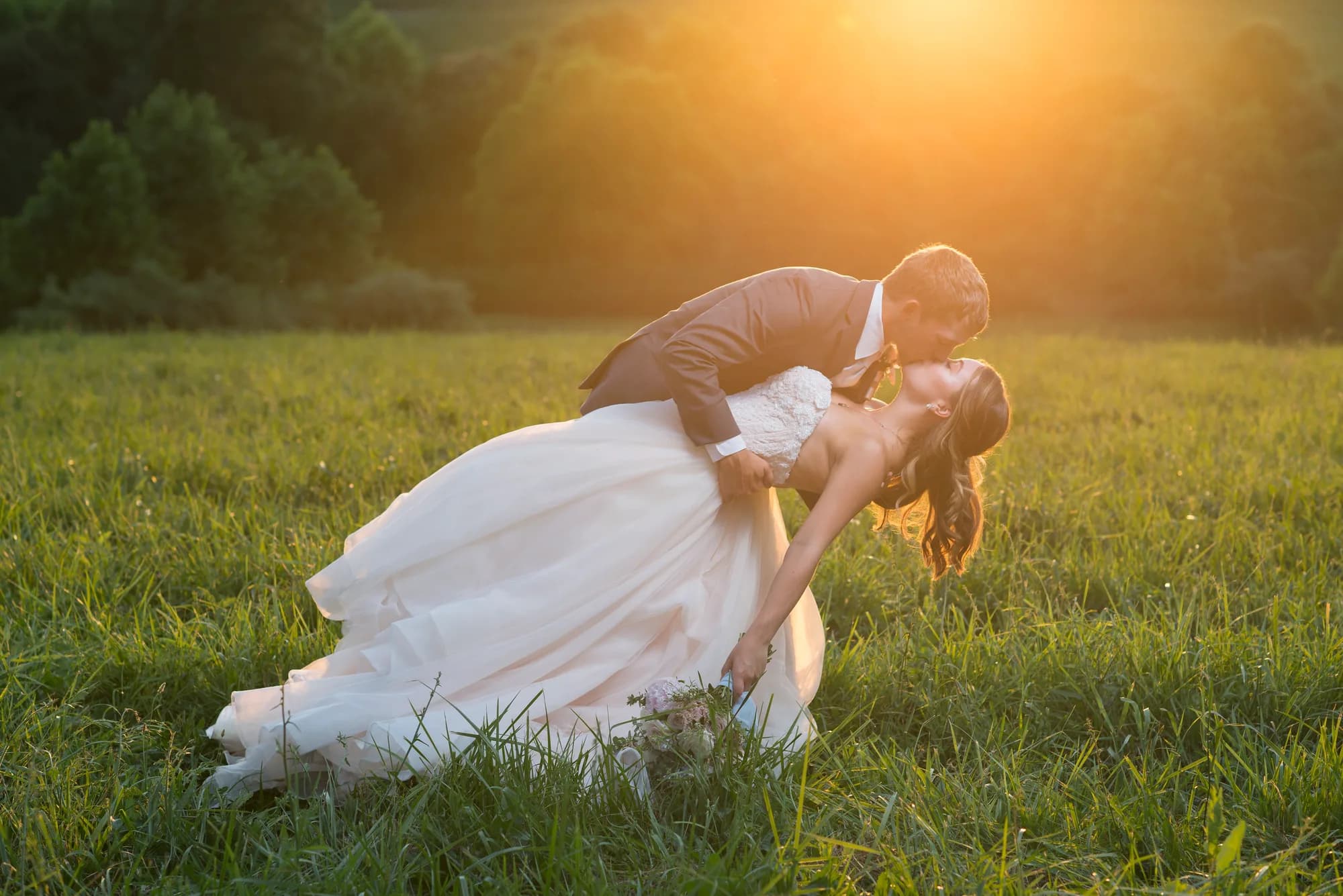 Bride and groom share a golden-hour dip kiss in a lush green field at sunset