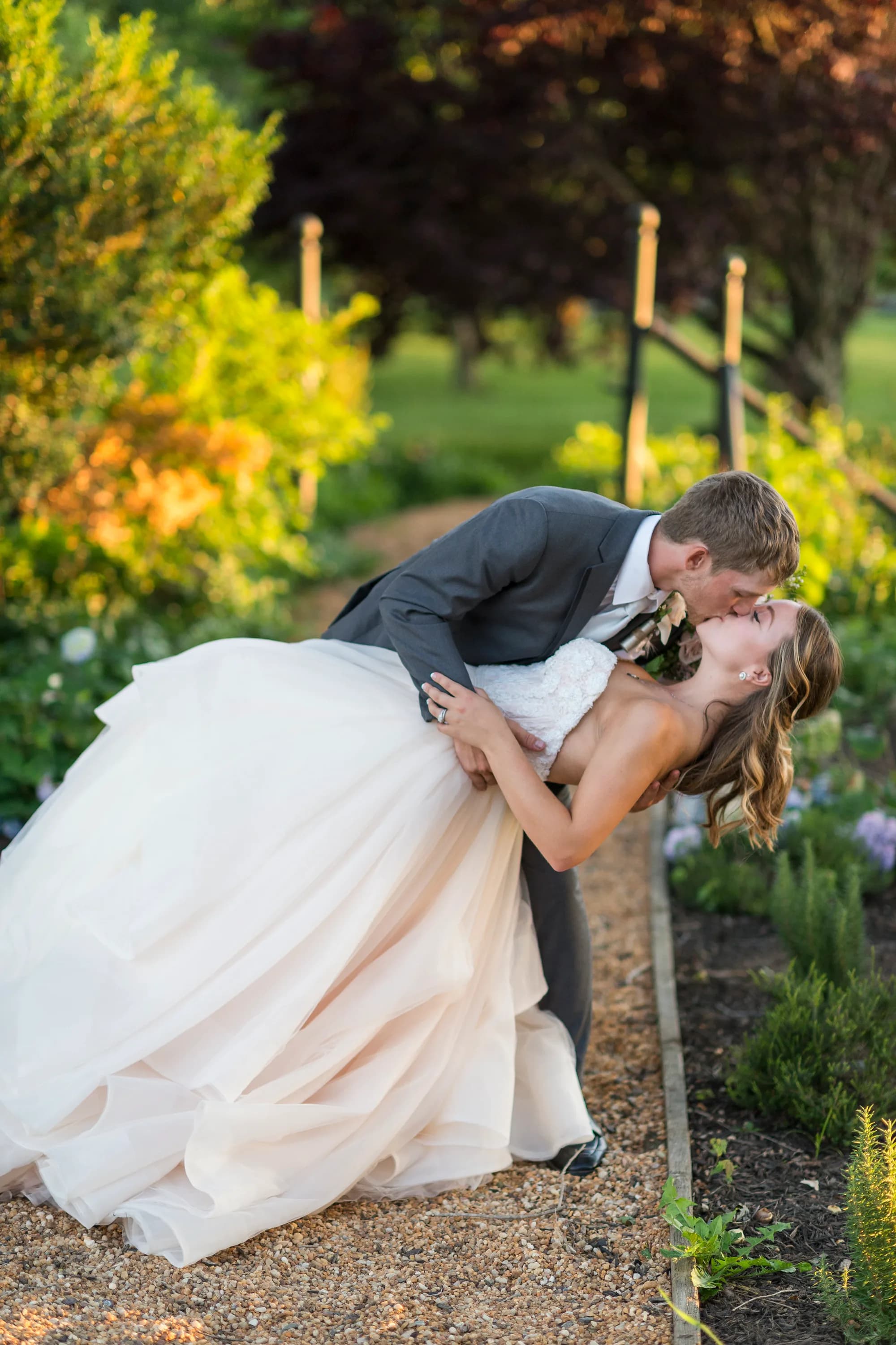 Groom dips bride for a kiss along a sunlit garden path at Rixey Manor, surrounded by lush greenery