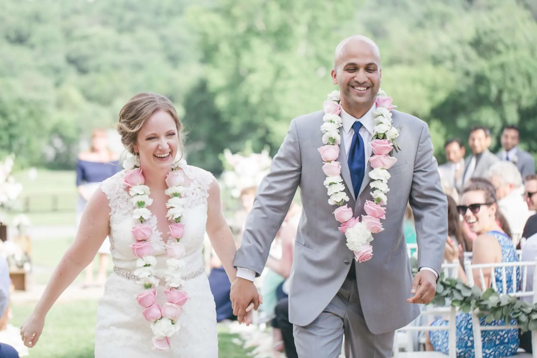 Laughing newlyweds wearing floral leis walk back down the aisle at an outdoor Virginia wedding ceremony