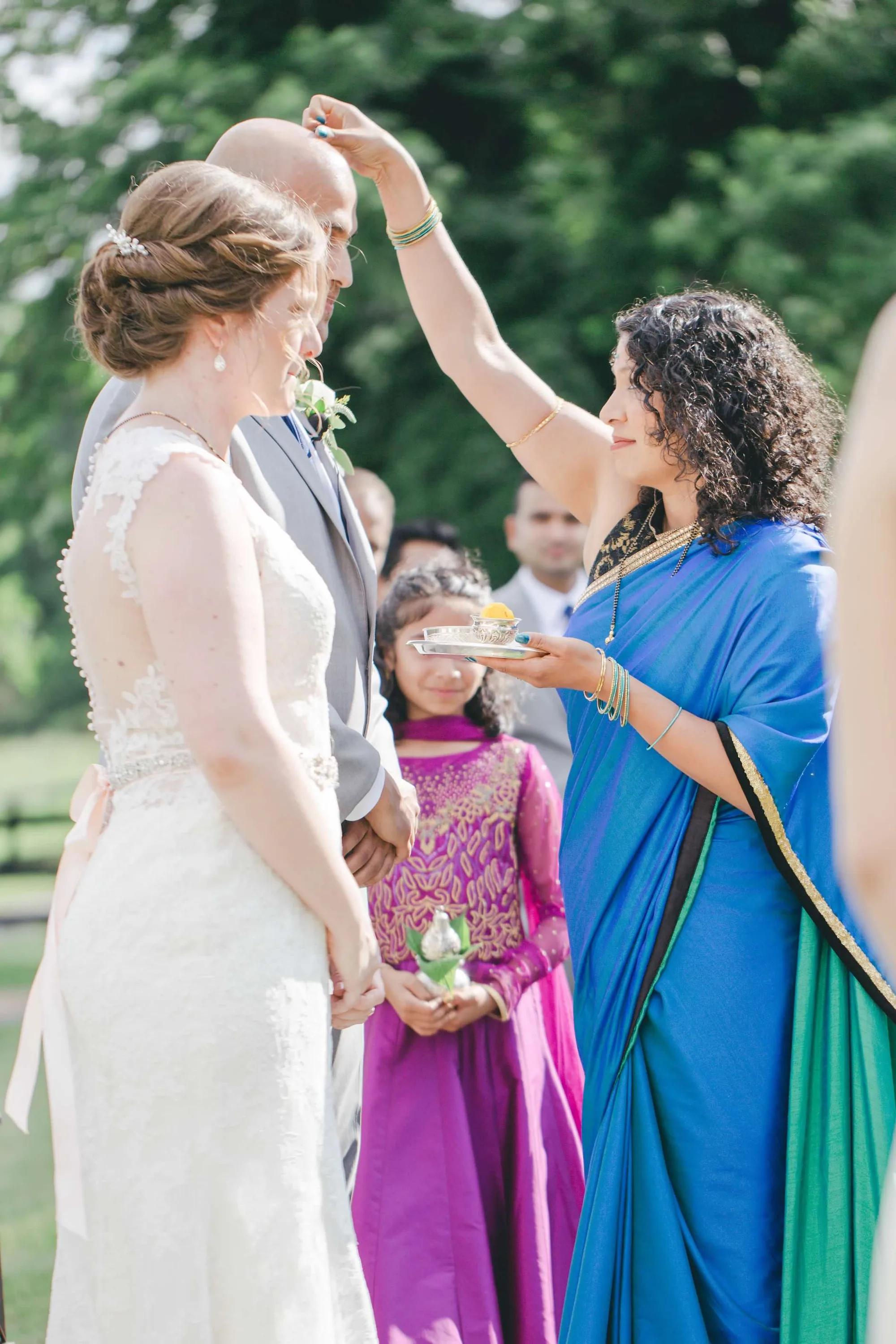Woman in blue sari performs Indian blessing ritual on couple during outdoor wedding ceremony