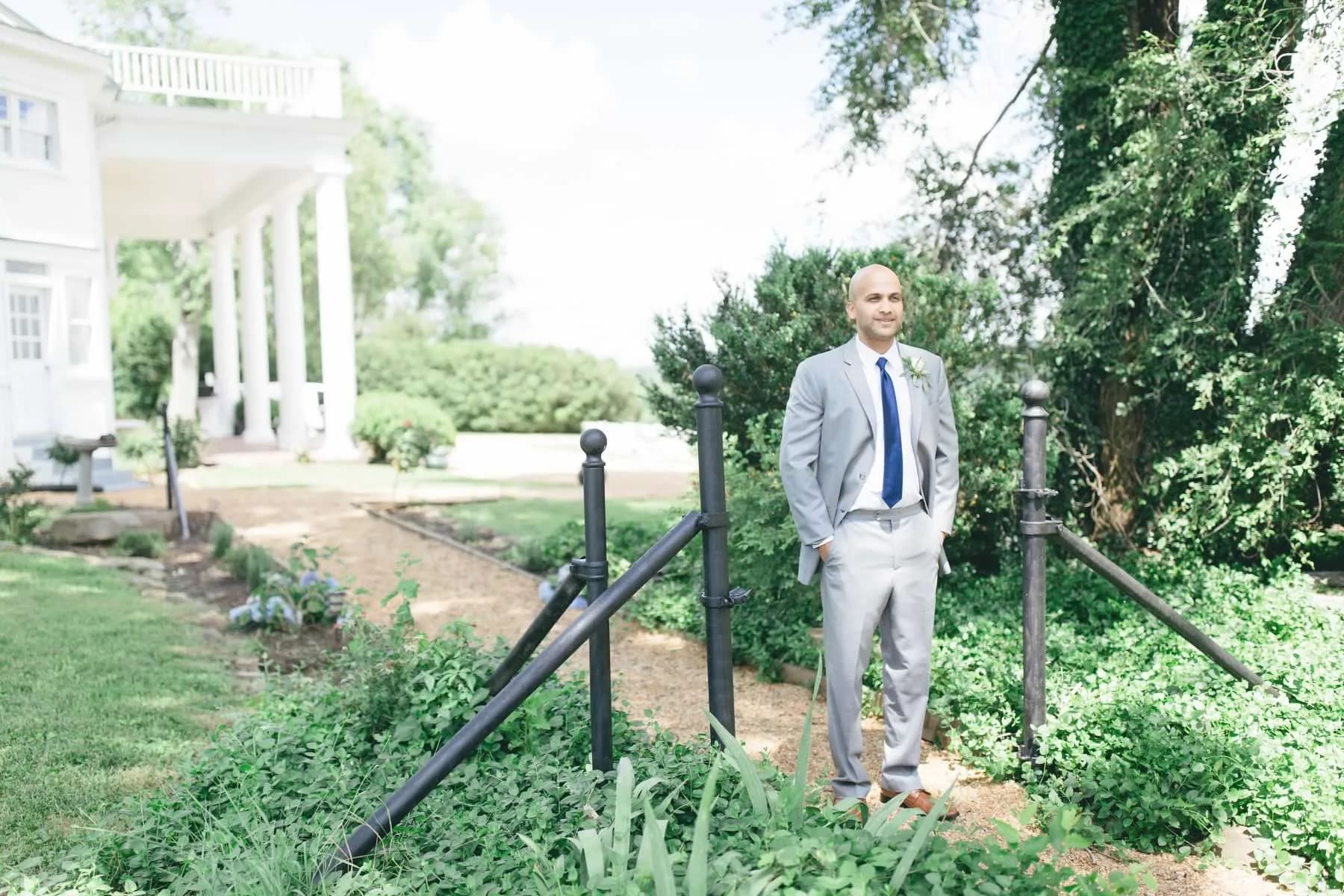 Groom in grey suit stands near iron fence amid lush gardens beside Rixey Manor's white columned estate
