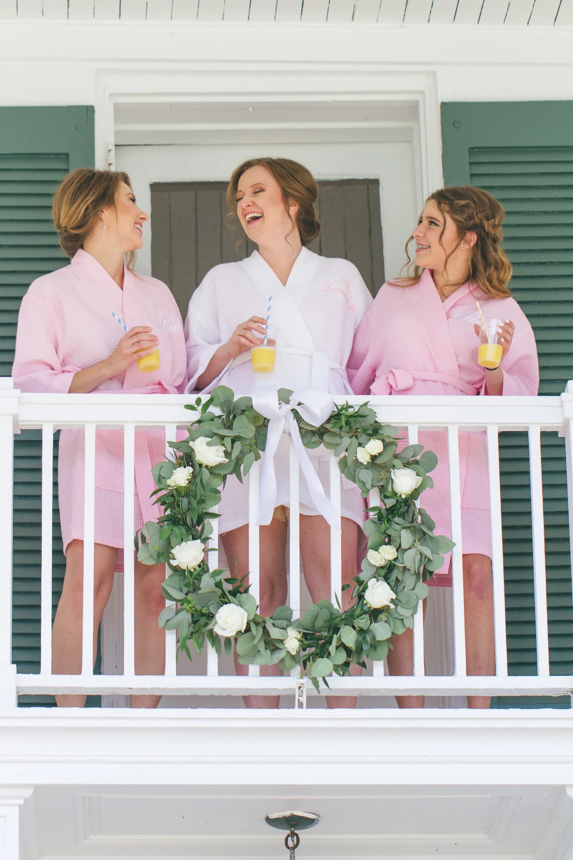 Bride in white robe laughing with bridesmaids in pink on a balcony decorated with a eucalyptus wreath