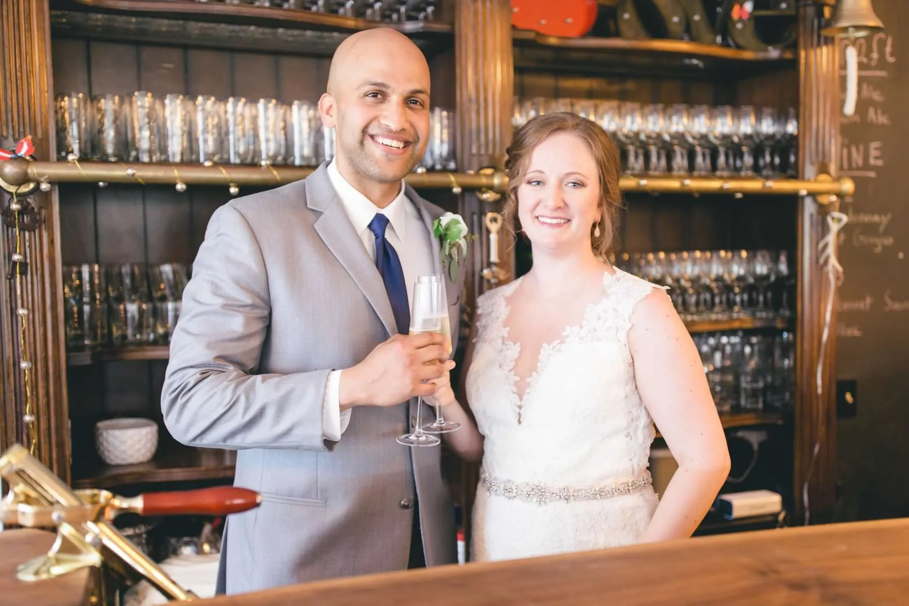 Bride and groom toast with champagne flutes at a wood-paneled bar, smiling warmly at the camera.