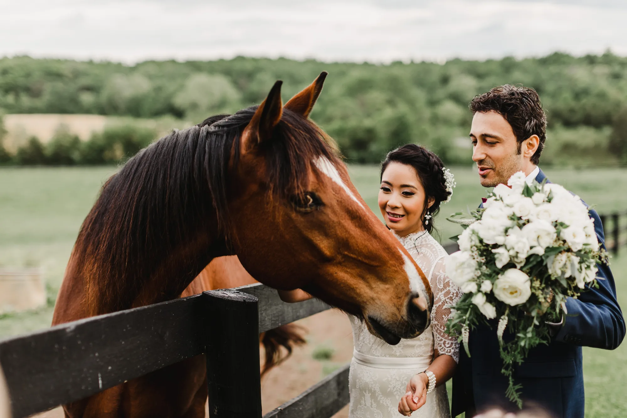 Bride and groom smiling as a chestnut horse nuzzles their white bouquet by a fence at a rural estate