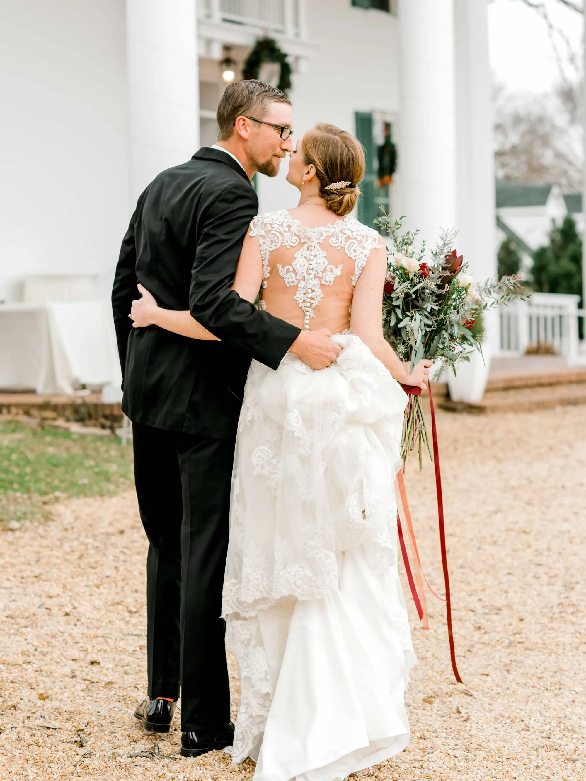 Bride and groom share a kiss outside Rixey Manor's white columned facade, bride holding red and greenery bouquet