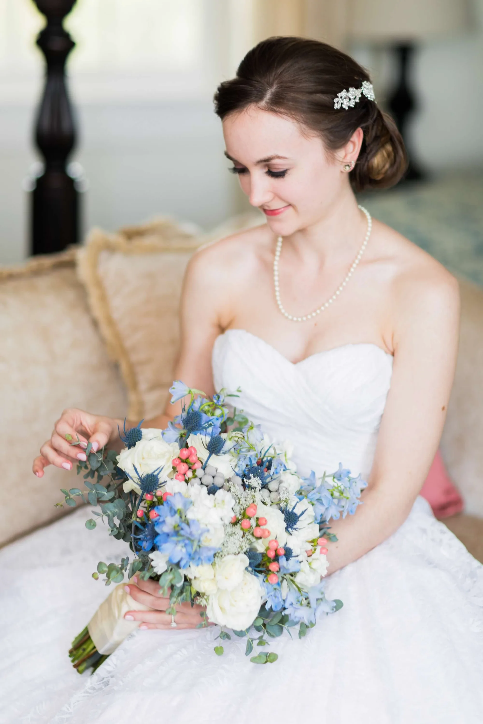 Bride in strapless gown and pearl necklace gazes at her bouquet of blue, white, and coral flowers