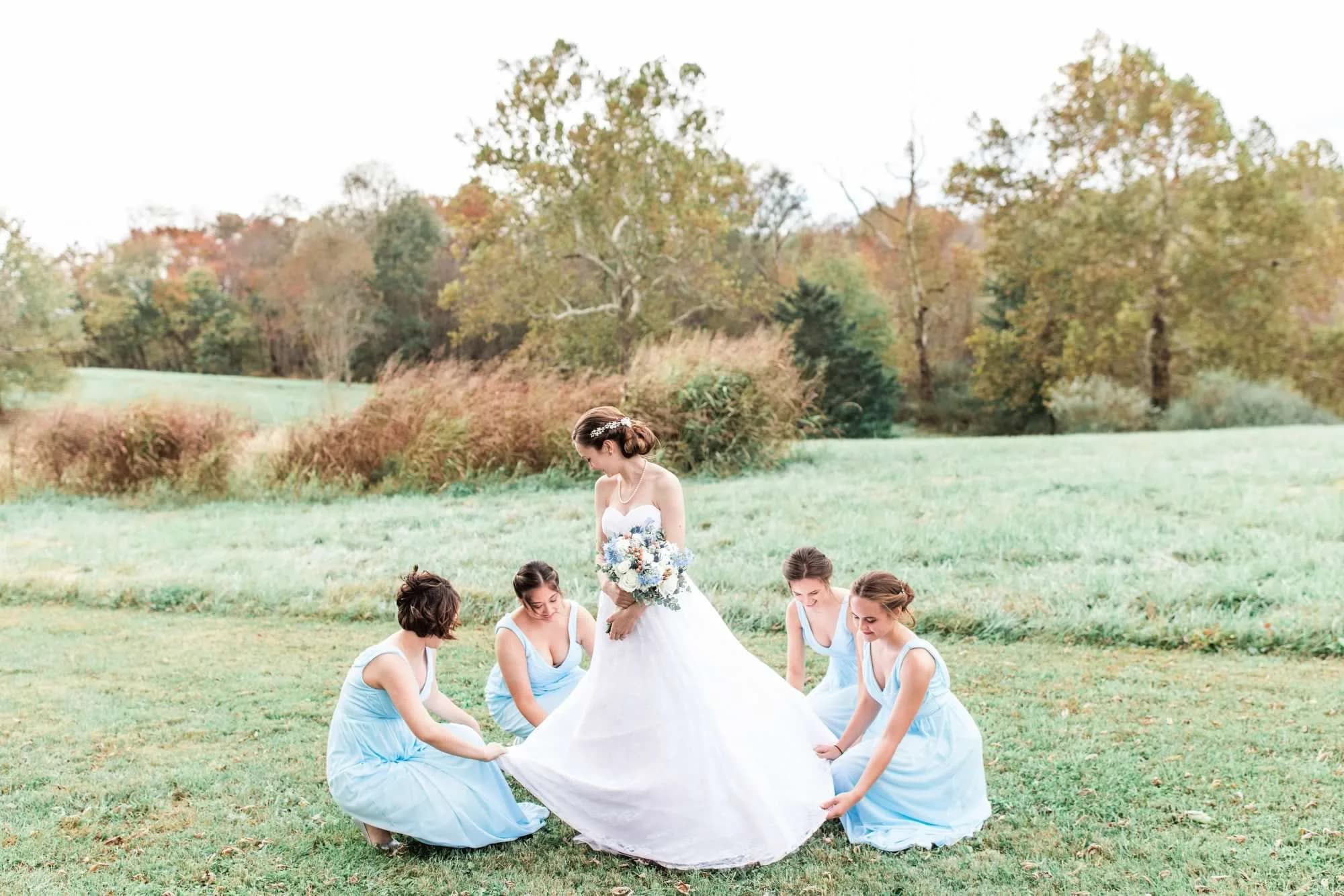 Bride in white gown holds bouquet as four bridesmaids in blue arrange her dress on Rixey Manor's green lawn
