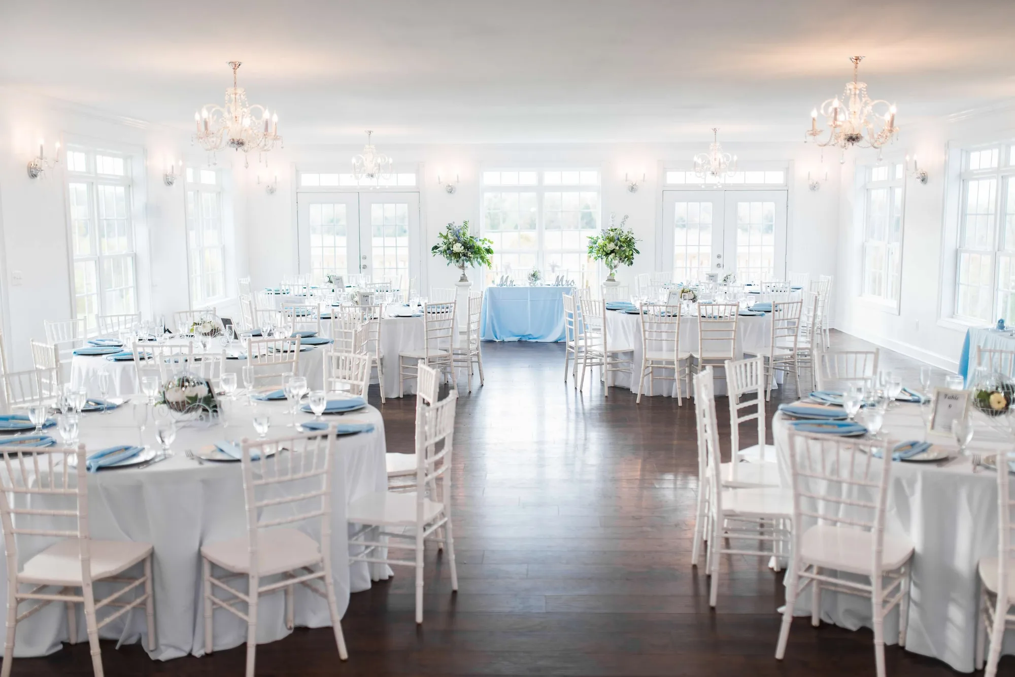 Elegant white reception hall with round tables, blue accents, chiavari chairs, and chandeliers
