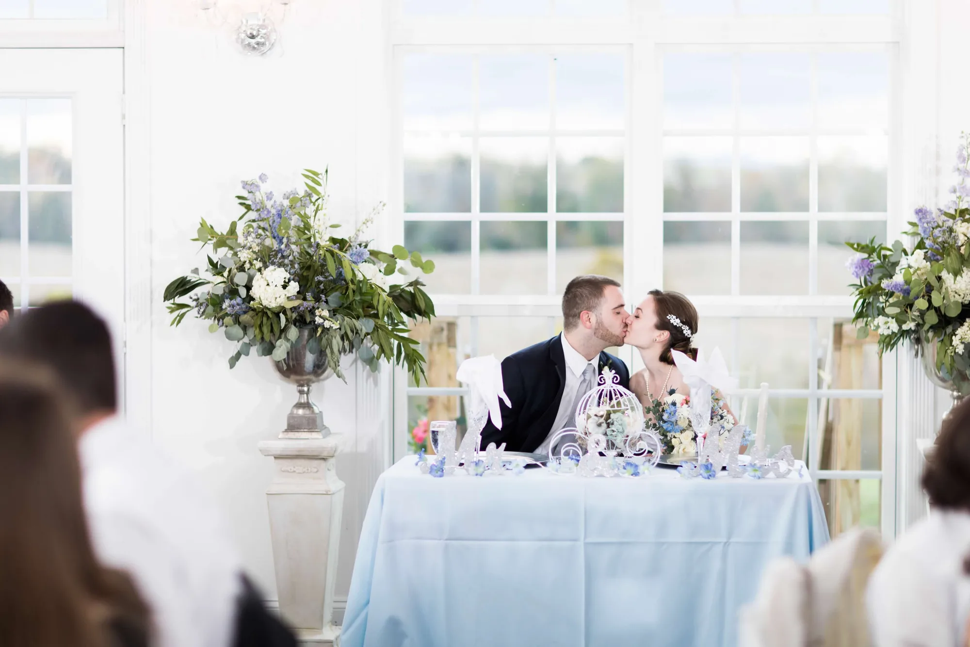 Bride and groom share a kiss at their sweetheart table in a bright, airy reception hall with floral arrangements