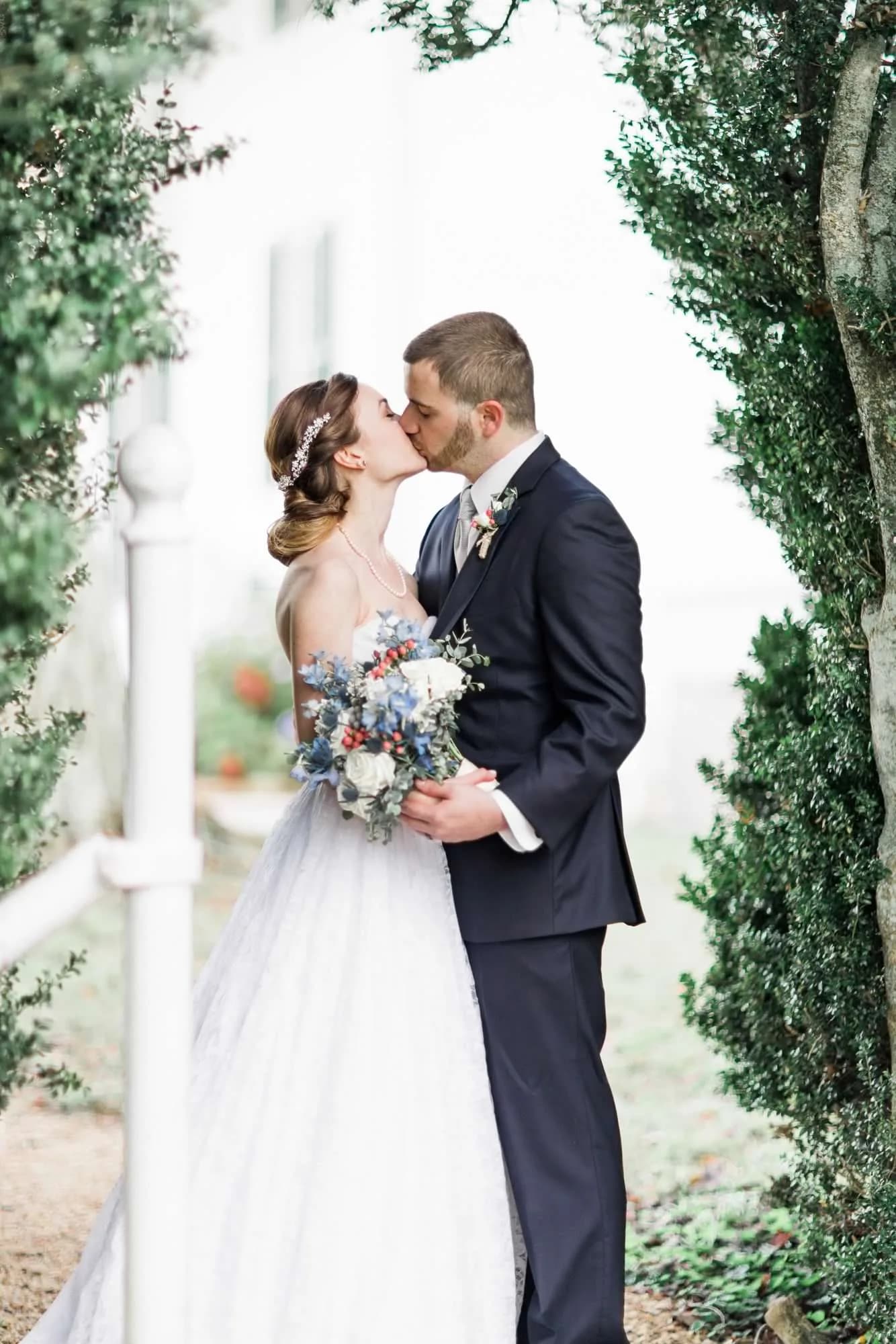 Bride and groom share a kiss framed by lush hedges at Rixey Manor, bride holding blue and white bouquet