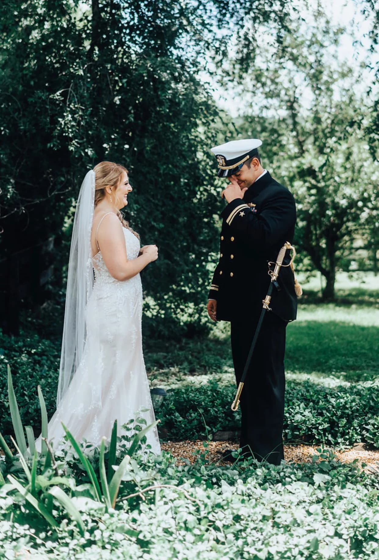 Groom in Navy dress uniform wipes tears at first look while bride smiles in lush garden setting