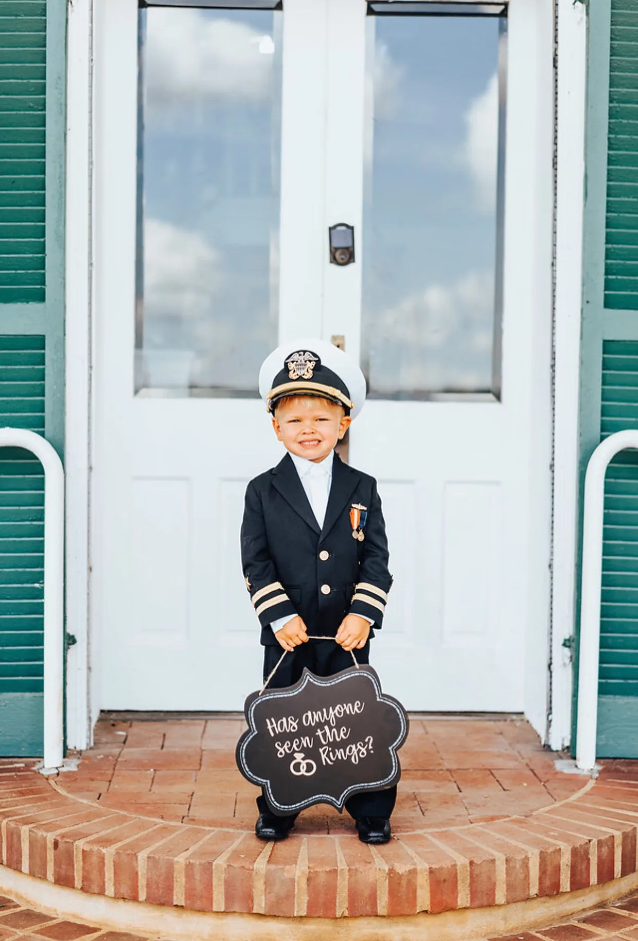 Smiling toddler ring bearer in naval uniform holds chalkboard sign reading 'Has anyone seen the rings?'
