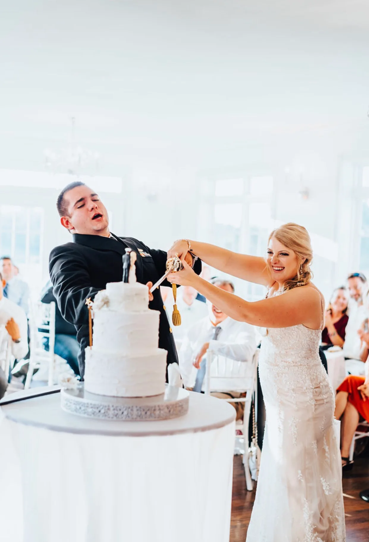 Bride and groom cutting white wedding cake together at reception with guests in background