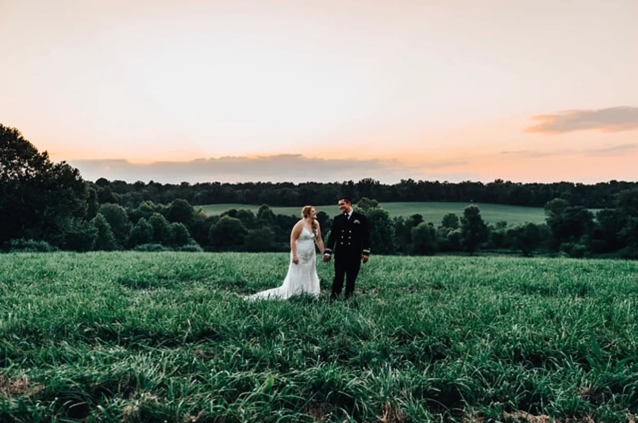Bride and groom standing in a green field at sunset with trees and sky in background