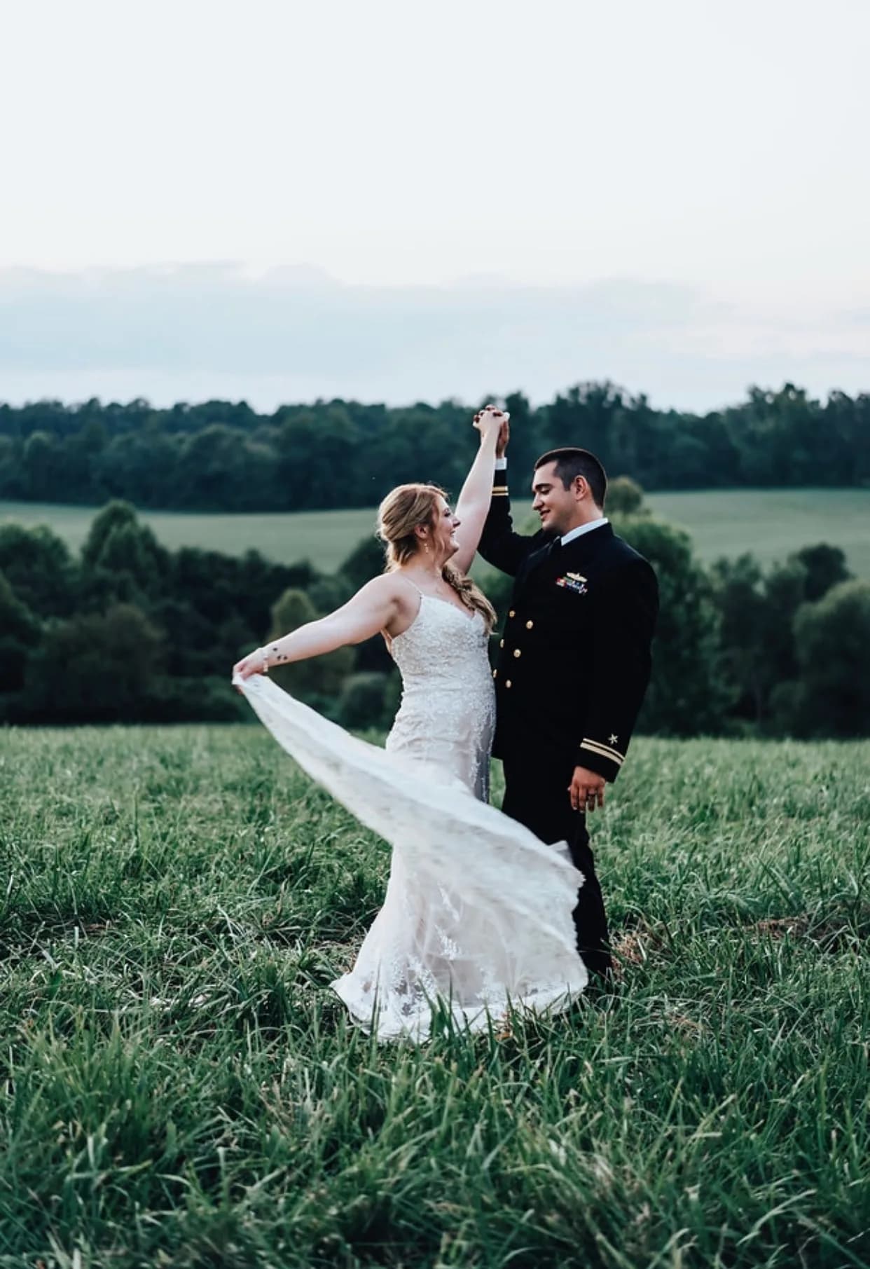 Bride in lace gown twirls with Navy groom in uniform across lush green fields at Rixey Manor