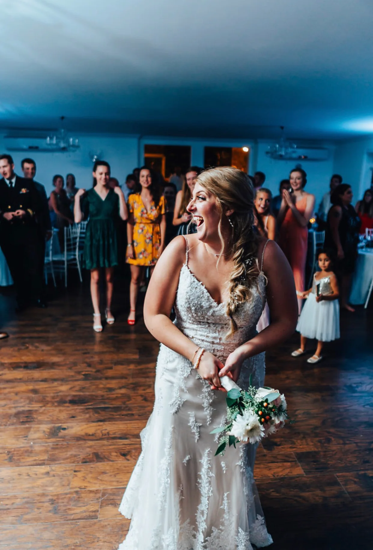 Bride laughing mid-bouquet toss on the dance floor as wedding guests look on in anticipation