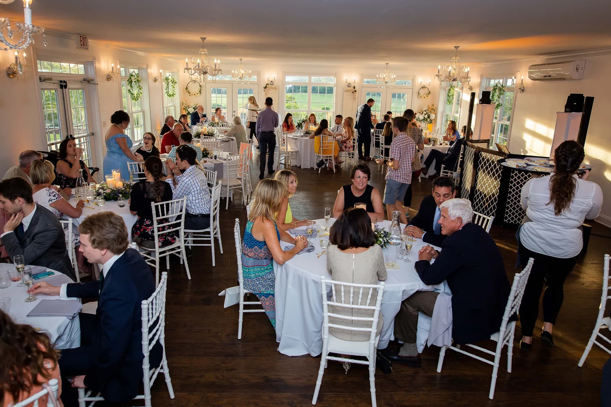 Guests seated at round tables during wedding reception inside Rixey Manor ballroom with white chairs and chandeliers