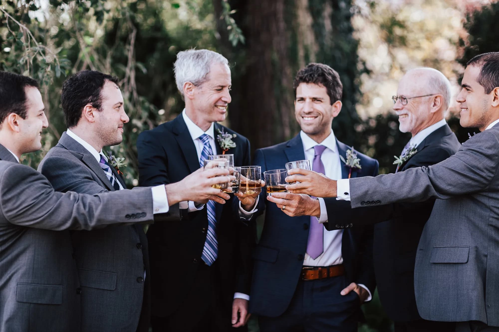 Six groomsmen in suits toasting with whiskey glasses outdoors, laughing together before the wedding ceremony