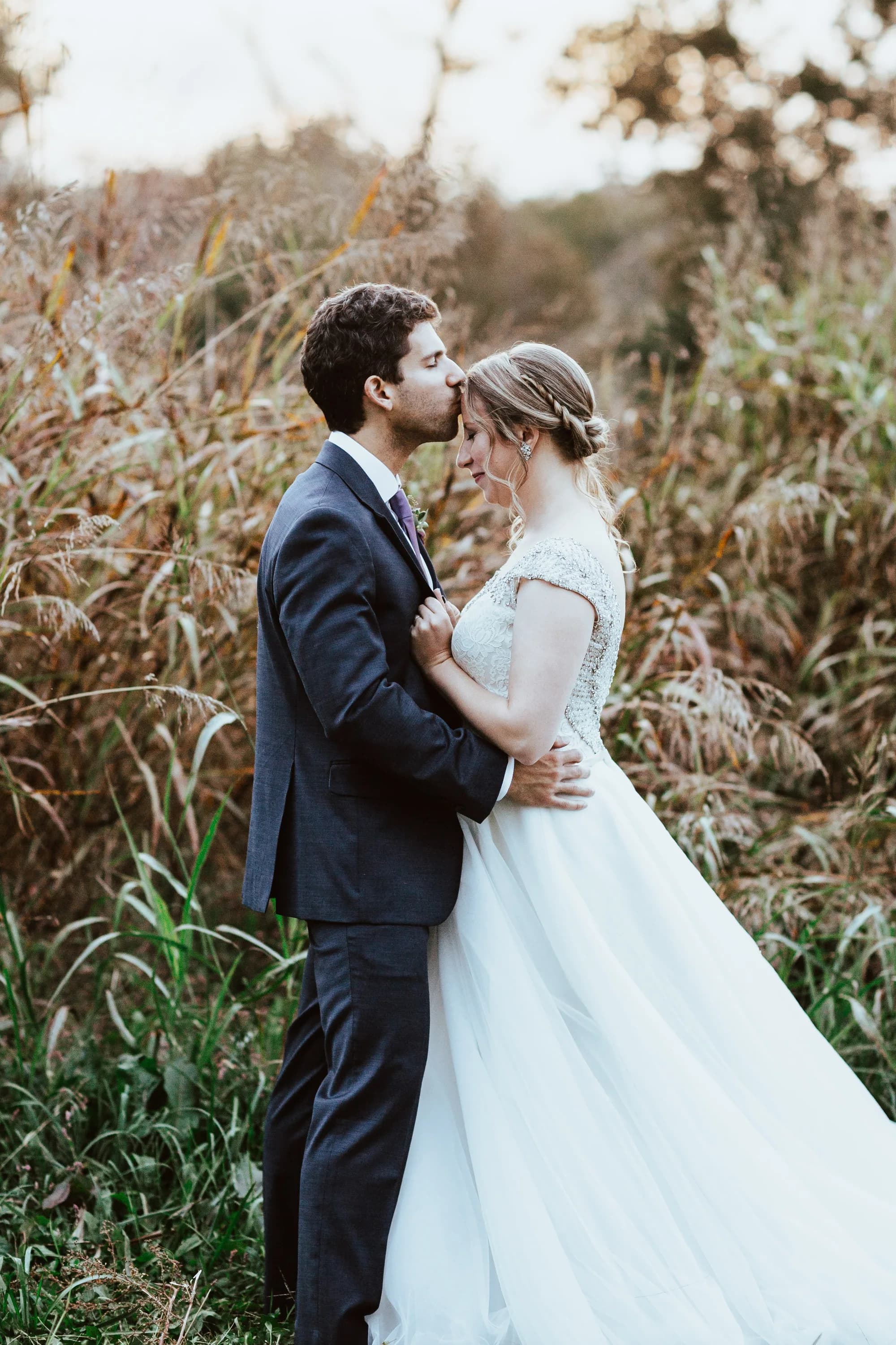 Groom kisses bride's forehead among golden autumn grasses during sunset portraits