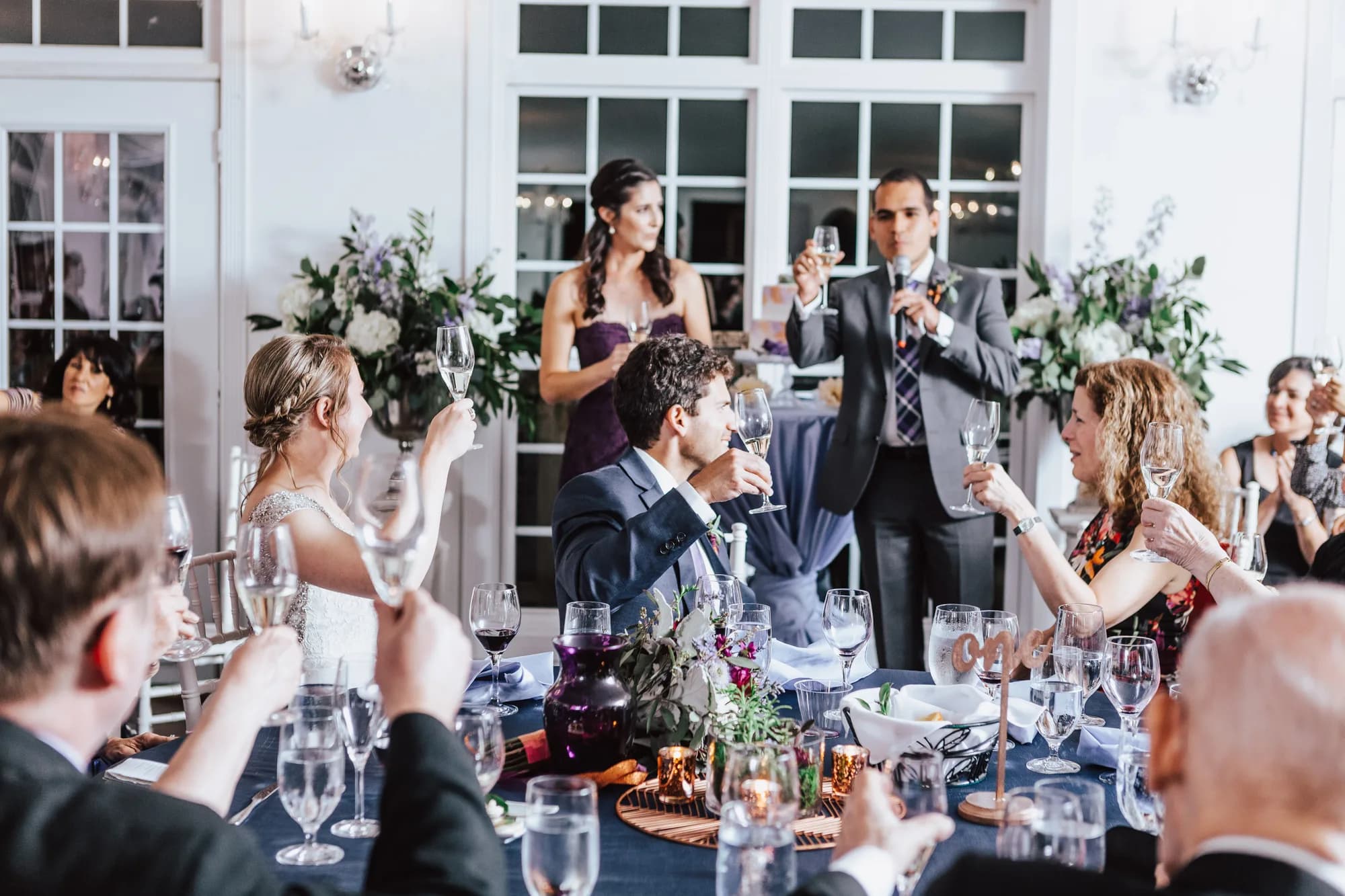 Wedding guests raise champagne glasses in a toast at the reception dinner table as the couple drinks