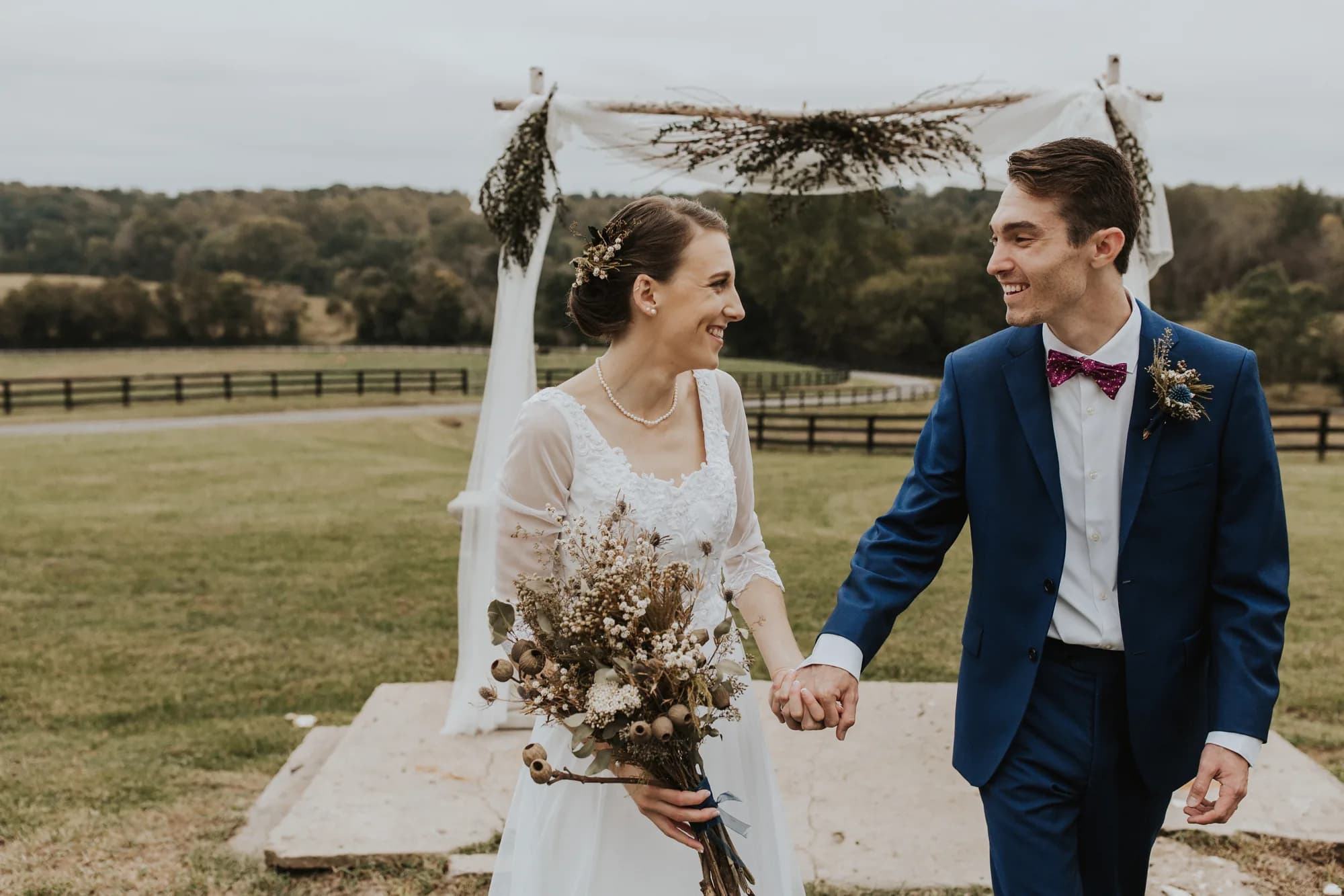 Smiling newlyweds hold hands walking from outdoor ceremony arch on Rixey Manor's pastoral grounds