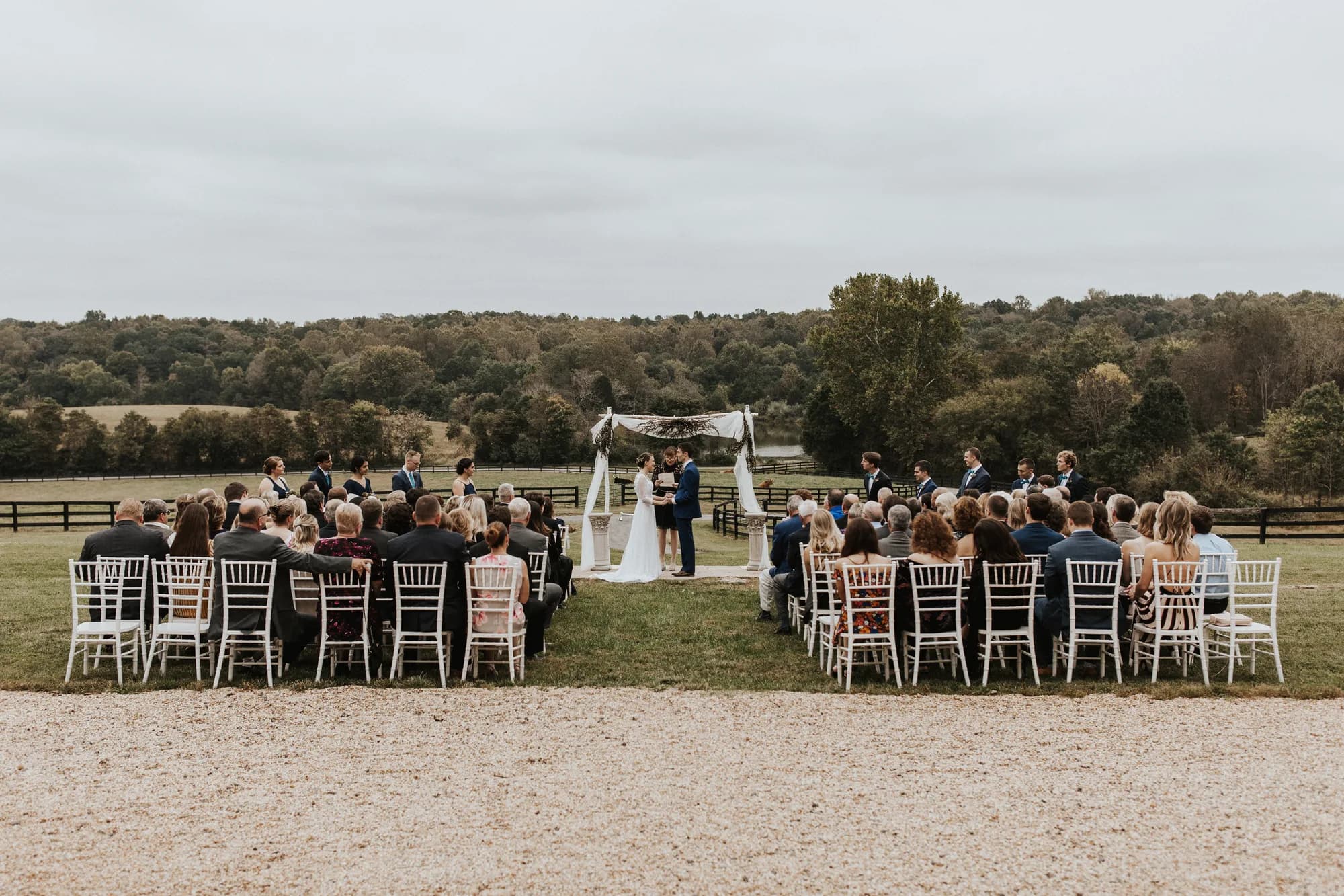 Wide shot of outdoor wedding ceremony on Virginia estate grounds with guests seated in white chairs facing couple at altar