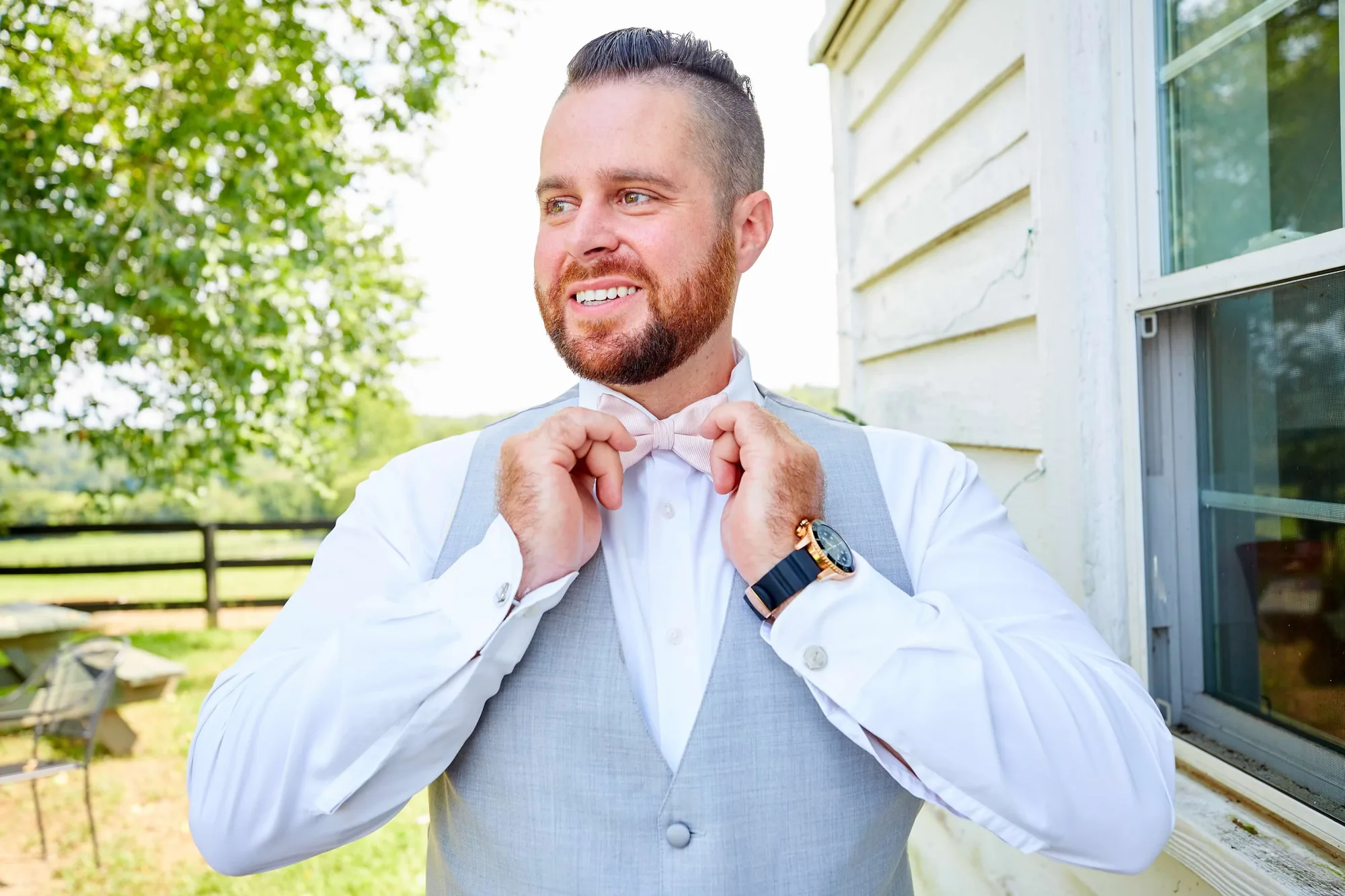 Smiling groom adjusts pink bow tie outdoors in gray vest and white shirt before his wedding ceremony