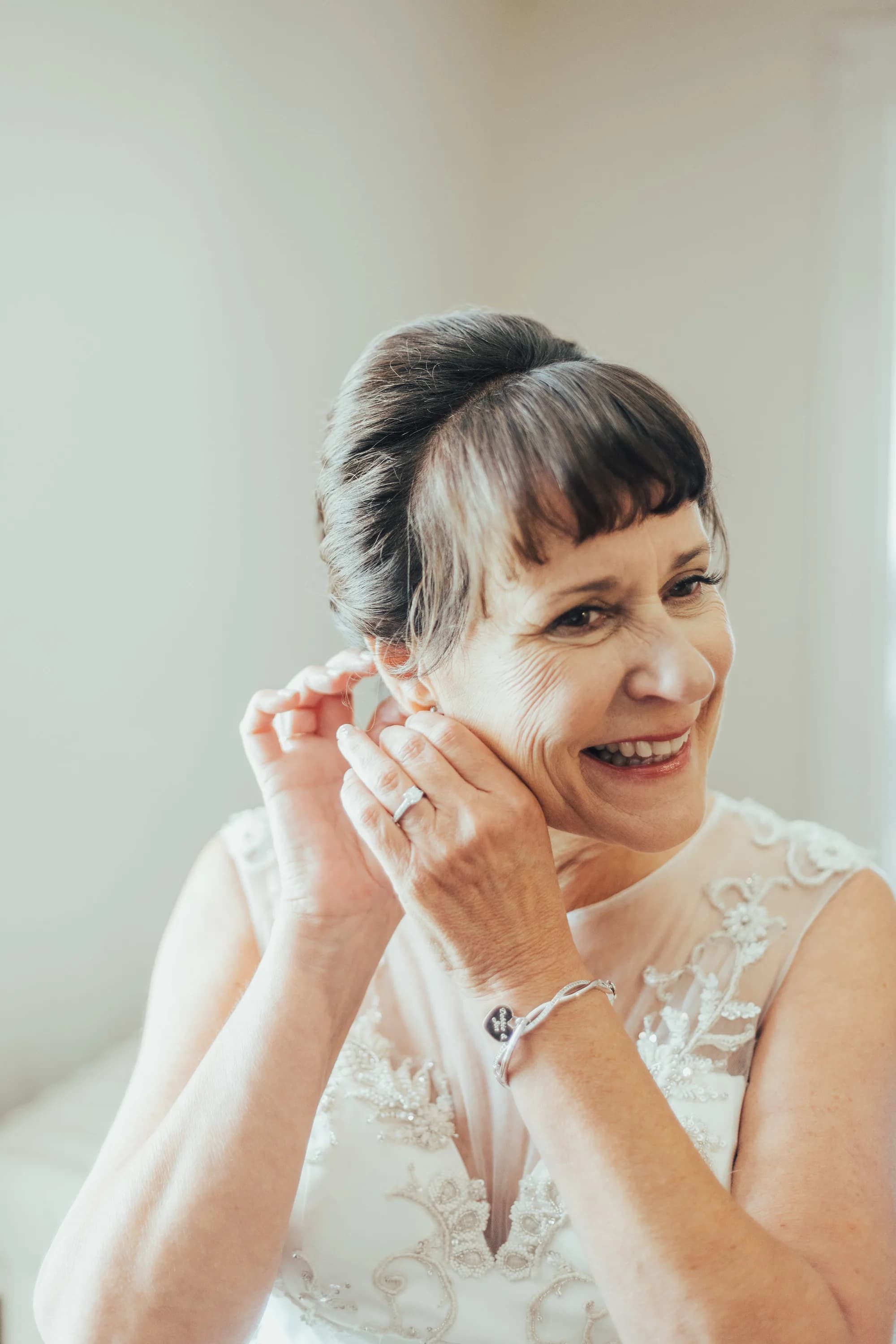 Smiling bride puts on pearl earring while getting ready, wearing lace wedding gown and diamond ring